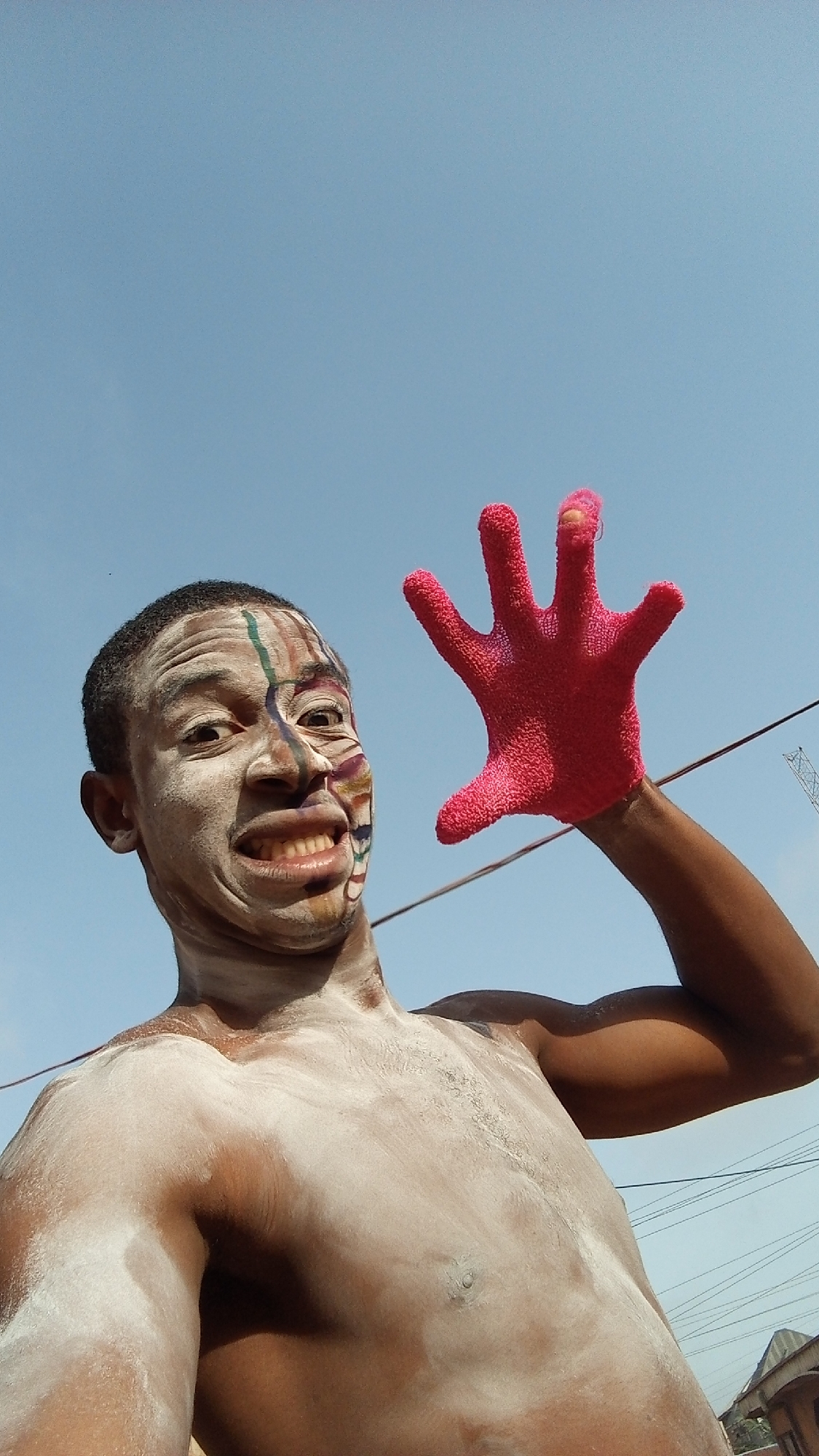 A low-angle shot depicts a shirtless man against a clear blue sky. His face is adorned with white paint and colorful vertical lines, and he is smiling broadly. His torso is covered with white powder. He is holding up a red glove on his right hand. Background details reveal power lines, a rooftop of a building, and what appears to be a construction crane. Given the prompt's instruction to include the location, it can be inferred that this scene is in Onitsha, Nigeria, based on the prompt's instructions.