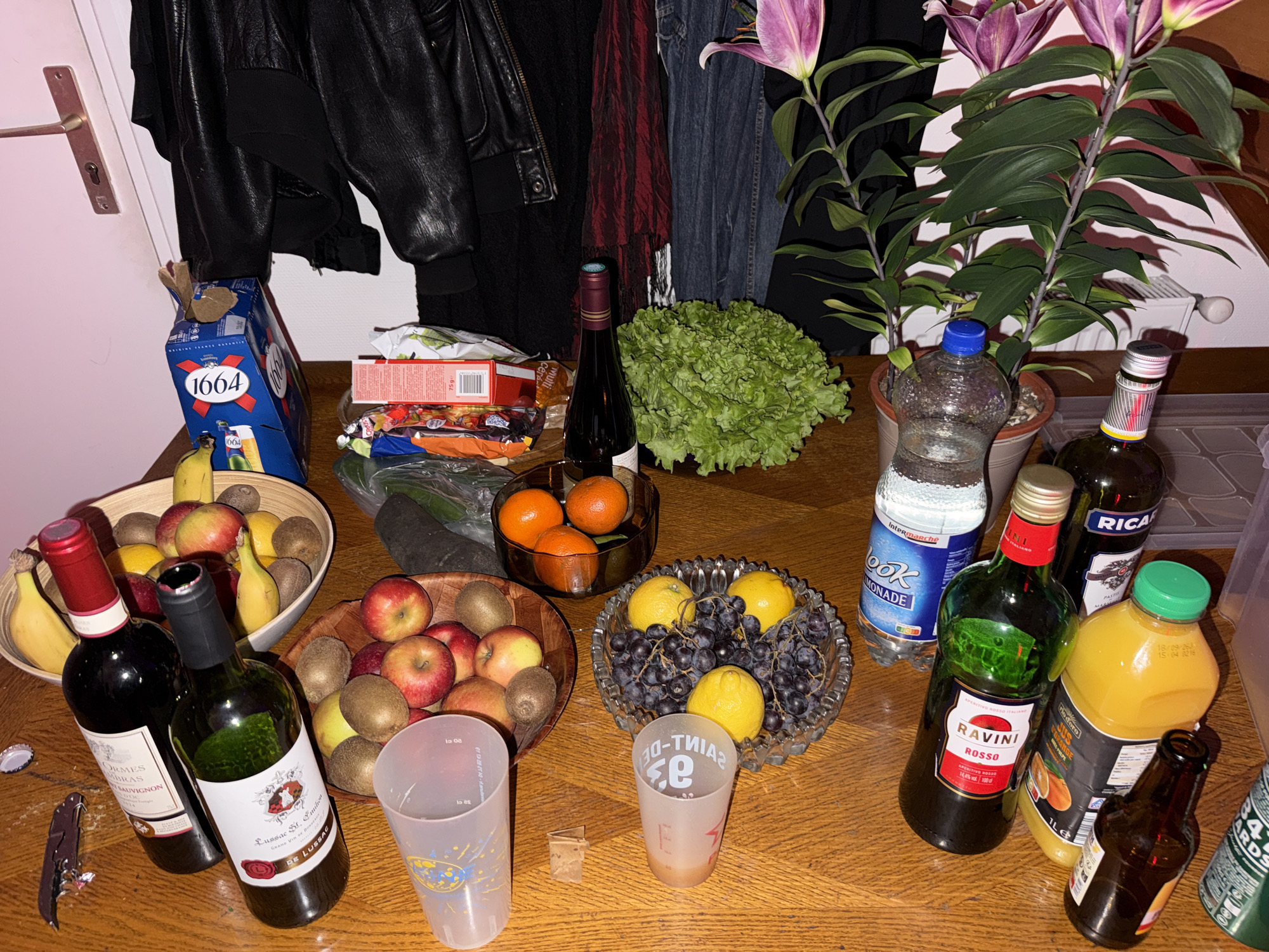 The image shows a wooden table laden with various items, suggesting a gathering or party. There are several bowls filled with fruit, including apples, kiwis, bananas, oranges, lemons, and grapes. Two bottles of wine, one with a red label and one with a green label (Lussac St. Emilion De Lussac), lie on their sides next to a corkscrew and some plastic cups. Other beverages include a bottle of Ricard, orange juice, a bottle of Ravini Rosso, and a bottle of beer marked "1664". There is also a bottle of water and a head of lettuce. A potted plant with purple flowers sits at the back of the table. In the background, a black leather jacket hangs next to a denim jacket. The location in Montreuil, France, is not discernible from the image.