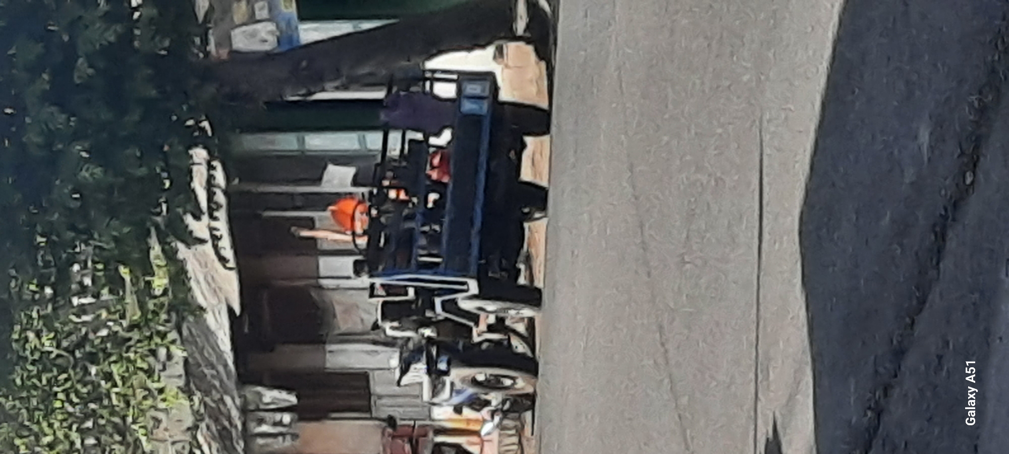 The image depicts a scene on a paved road in Selopuro, Indonesia, with a building and trees lining the left side of the road. In the foreground, the road surface is gray with visible cracks. A three-wheeled vehicle, blue in color with a flatbed cargo area, is parked or moving slowly on the right side of the road. The vehicle has a driver visible in the cab and a person wearing an orange helmet in the cargo area, along with several other figures or objects. A motorcycle is attached to the front of the three-wheeled vehicle. A large tree trunk is prominent on the right side of the image. The building in the background appears to be a low-rise structure with a corrugated metal roof and windows. Laundry is hanging from lines in front of the building. The overall impression is a snapshot of daily life on a street in Indonesia.