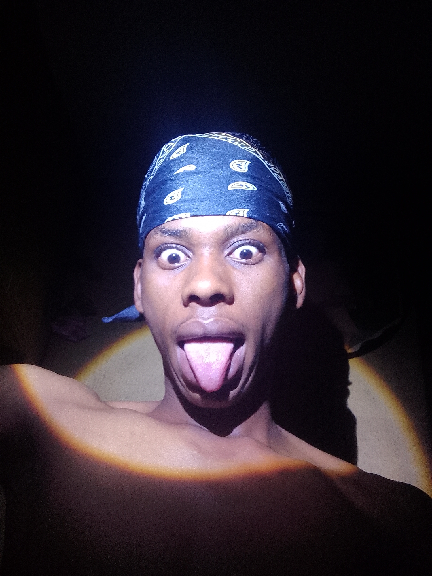 A close-up selfie of a shirtless Black male with a blue paisley bandana on his head. He is sticking his tongue out and has wide eyes. The background is dark. A light source casts a circular illumination on his left shoulder and chest. The location is Unknown, Unknown.