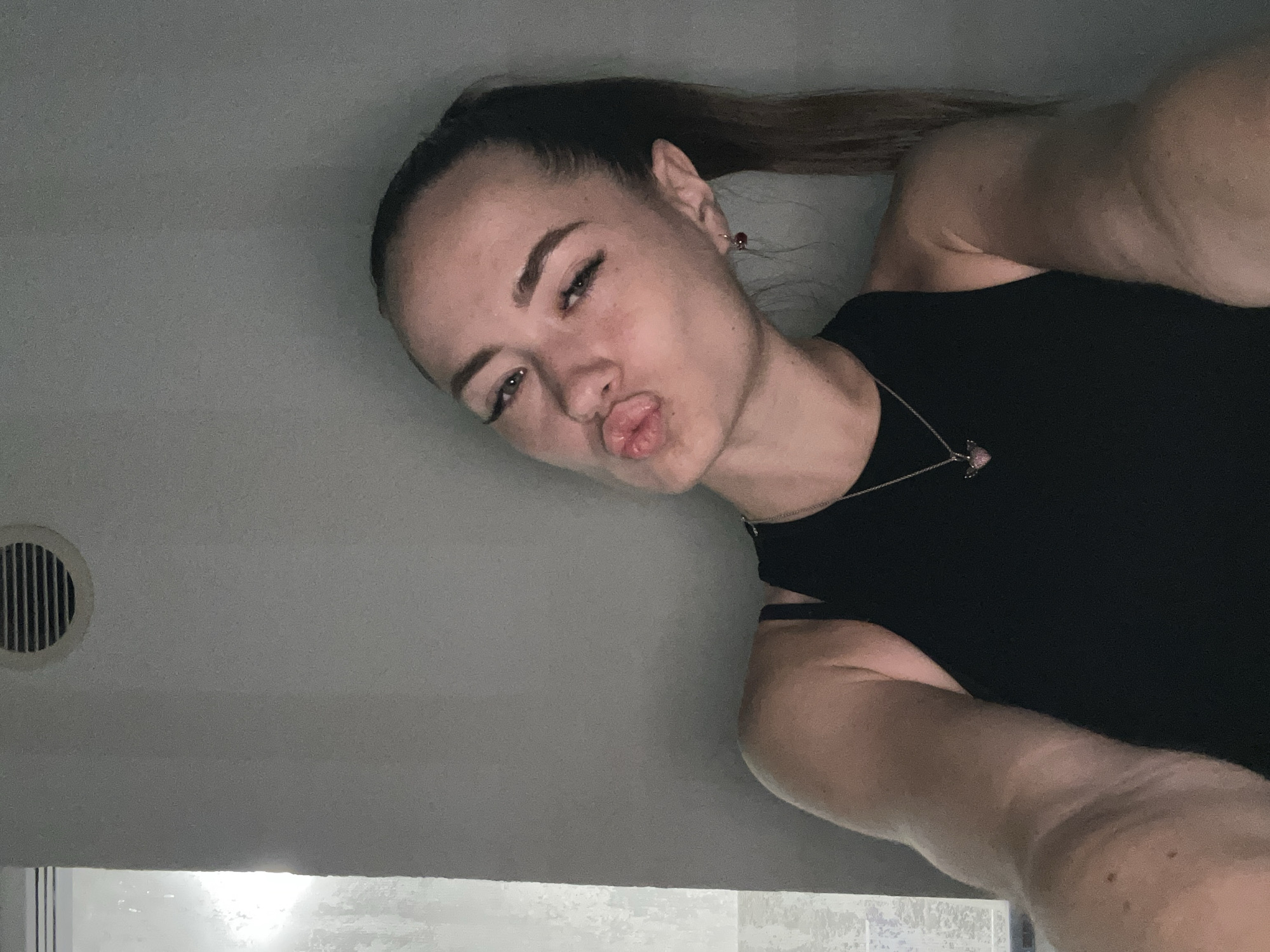 A young woman with her hair in a ponytail is taking a selfie against a plain wall. She is wearing a black sleeveless top and a necklace with a heart-shaped pendant. Her lips are pursed in a kissing gesture, and she is looking towards the camera. There is a circular vent on the wall behind her. The lighting is dim and casts a shadow on the wall. The overall scene is a casual, indoor setting.