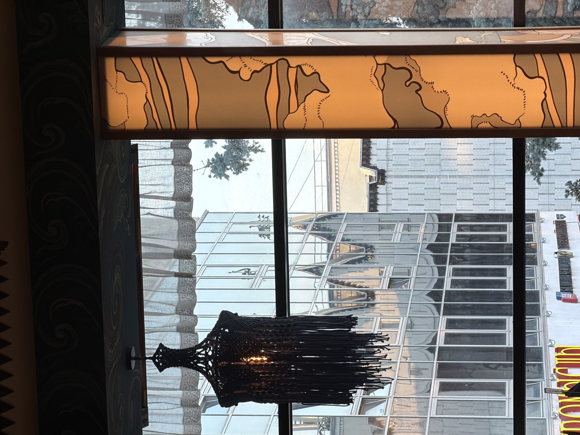 The image captures an interior view looking out through a window in Nizhny Novgorod, Russia. In the foreground, a macrame lampshade with a visible lightbulb hangs from the ceiling. To the right, a vertical art nouveau style lamp with an orange, glass panel featuring abstract line designs is illuminated. Beyond the window, a modern glass-facade building reflects the sky and the opposite architecture, showing a gothic-inspired structure. Further down, there is a glimpse of yellow signage with red lettering, suggesting commercial activity at street level. The overall atmosphere is dimly lit and artistic.