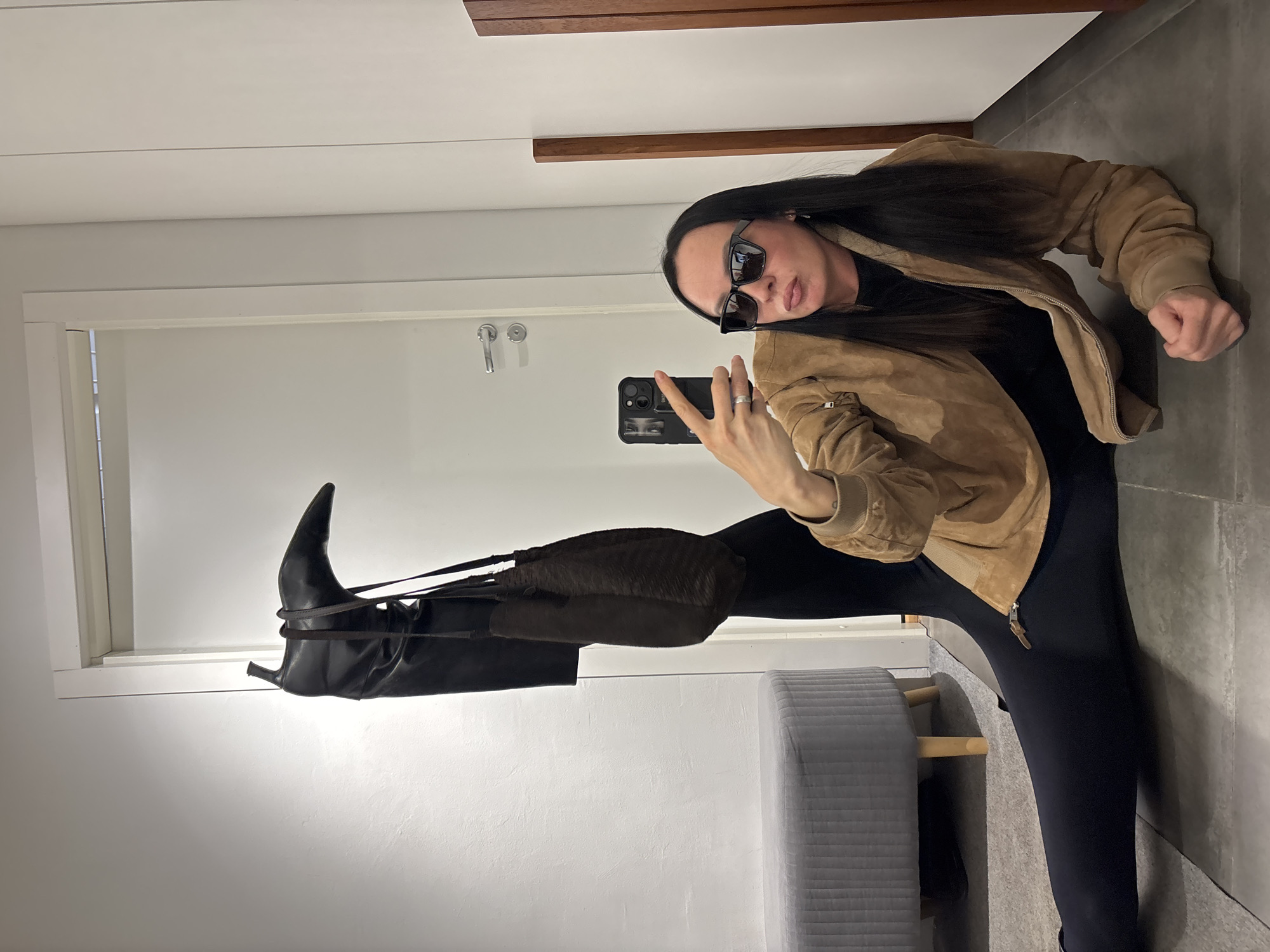 A woman takes a selfie in a fitting room. The woman is wearing sunglasses, a tan jacket, and black leggings. She is holding her leg up with a black knee-high boot and a brown purse on it. The background is a white fitting room with a gray ottoman and wooden details. The location is unknown and cannot be determined from the image.