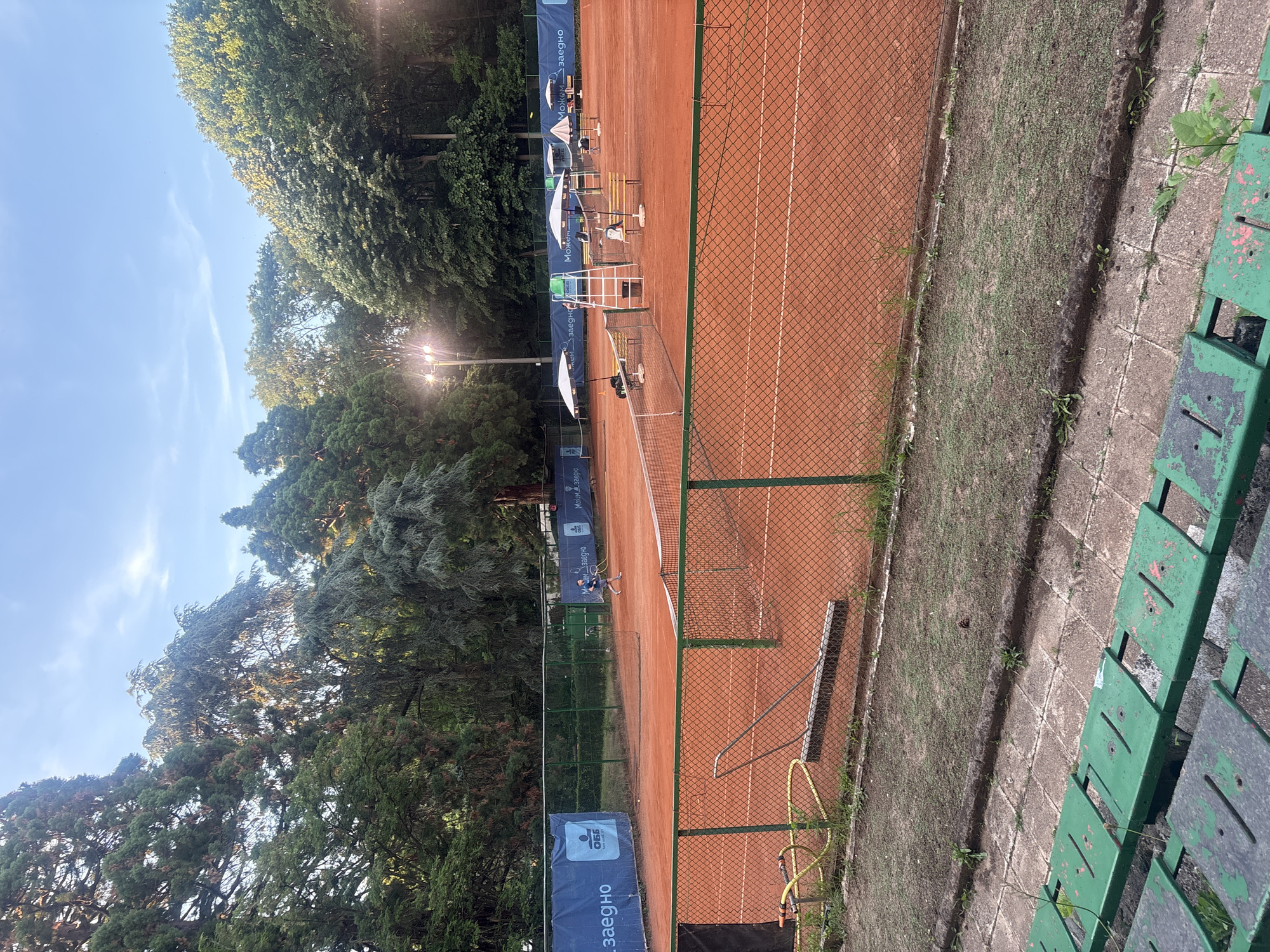 The image depicts an outdoor tennis court in Sofia, Bulgaria. The court is made of red clay and is surrounded by a green fence. In the background, there are tall trees, some of which are illuminated by artificial lights, suggesting it may be evening. A person is visible on the court, appearing to play tennis. There are also tennis nets, chairs, and umbrellas present. In the foreground, there are green bleachers, indicating spectator seating. The sky is blue with some clouds.