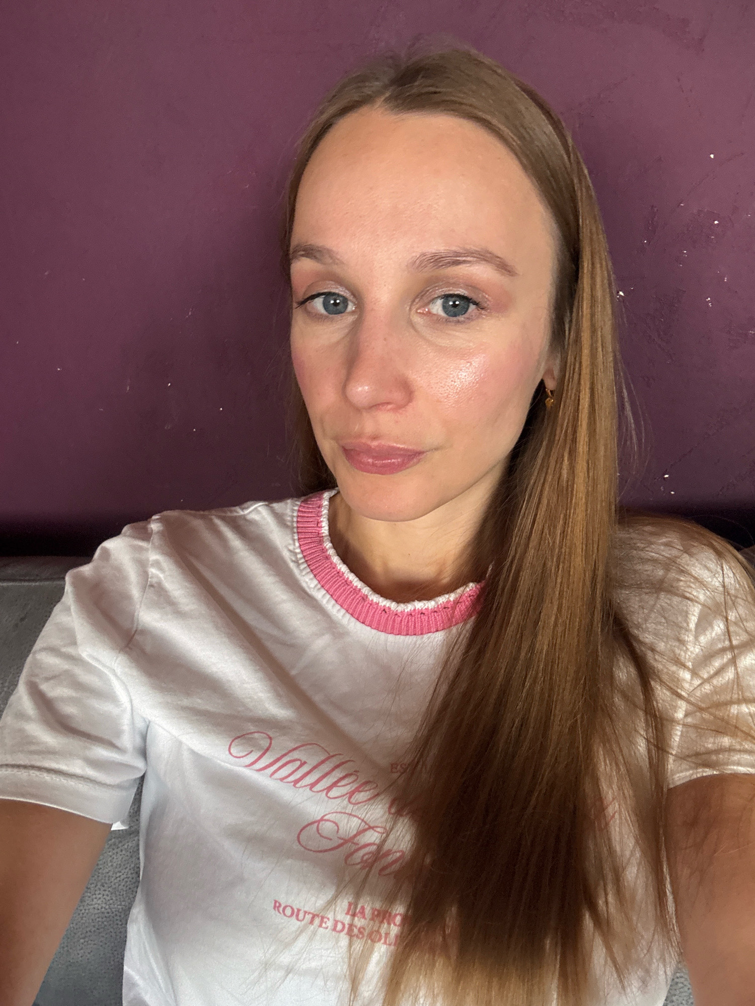 A selfie of a woman with long, blonde hair is shown against a purple wall in Überherrn, Germany. She is wearing a white t-shirt with pink trim and pink lettering that reads "Vallée des Fées" and "ROUTE DES OLIVES". The woman has blue eyes and light skin, and she is looking directly at the camera. The lighting in the image is soft and natural.