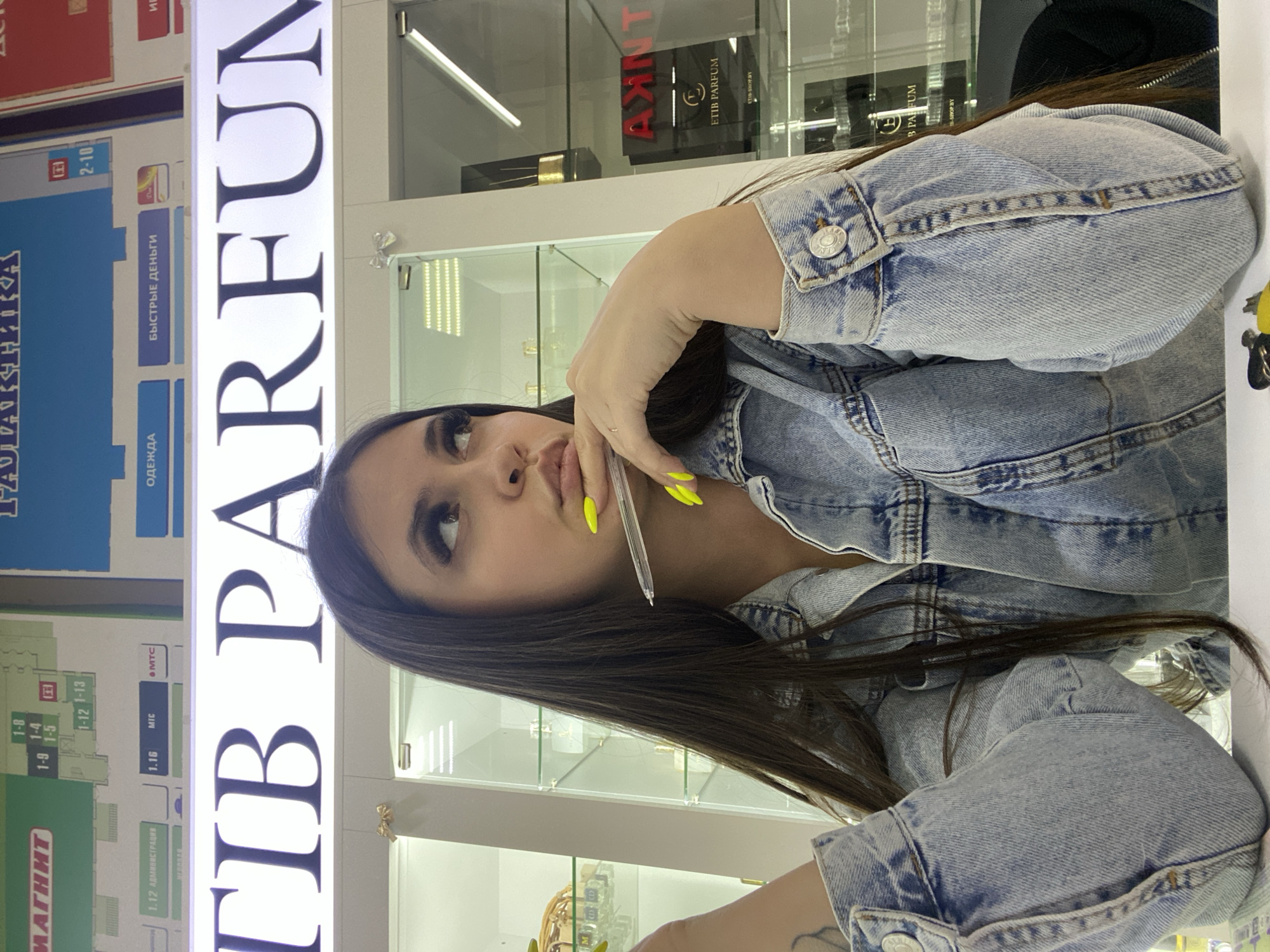 A woman with dark hair and vibrant yellow nail polish poses in front of a perfumery, "ПІВ PARFUM," likely within a shopping center, based on the directory signs. The location is the city of Chaikovsky, Russia. She is wearing a light blue denim jacket and holding a pen near her mouth. Behind her, shelves display various perfume brands, including "AXINT" and "ETIB PARFUM," as well as the online shop address. The directory includes store locations and categories like "Одежда" (Clothing), "Быстрые деньги" (Quick Money), and "Магнит" (a grocery store).