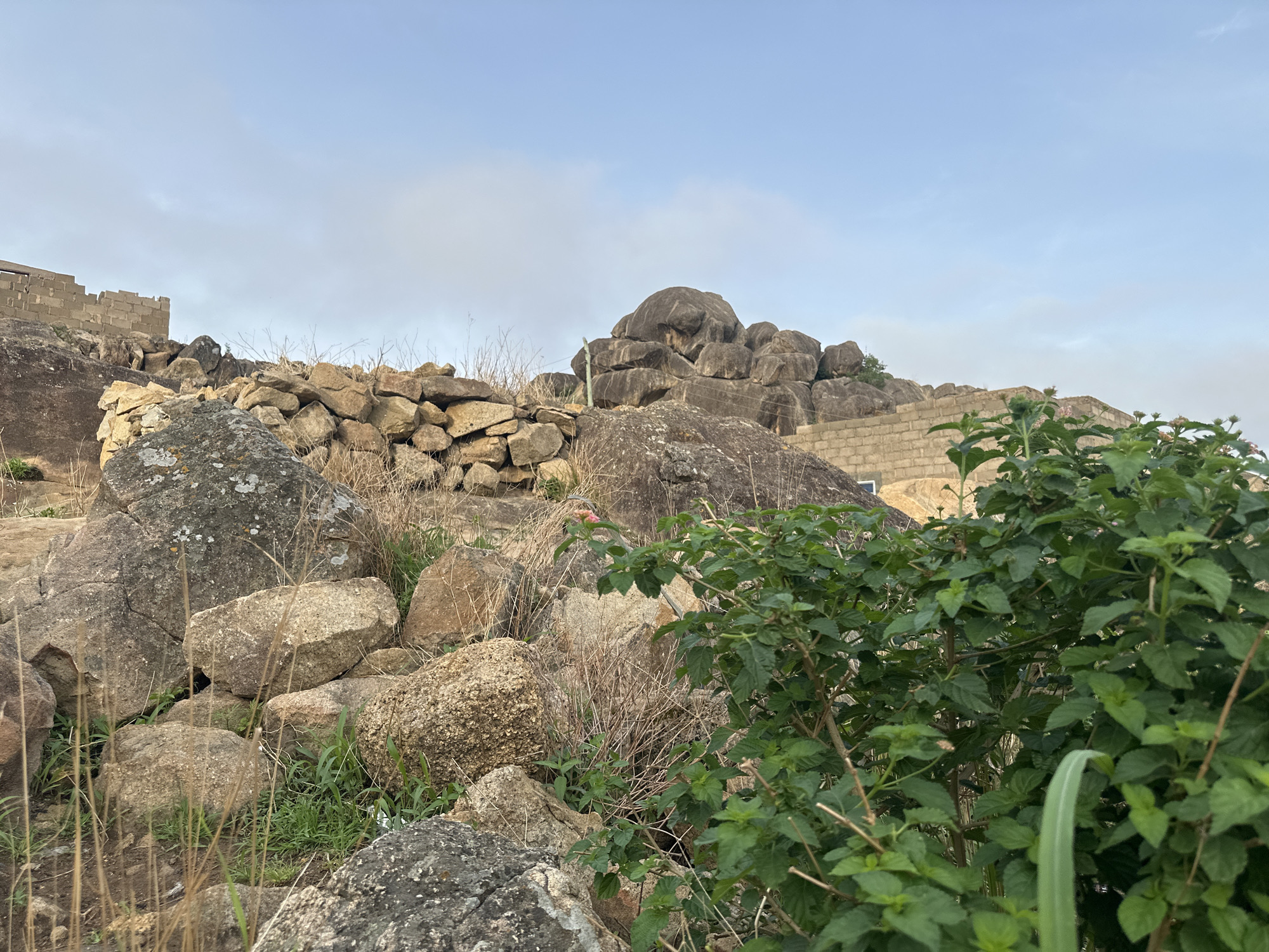 The image shows a rocky landscape with vegetation in Jos, Nigeria. The foreground includes rocks, dry grass, and a green bush. Further up the slope, there is a stone wall and a stack of large boulders. In the background, there is a concrete structure with an unfinished appearance. The sky is overcast. No people are visible in the image.