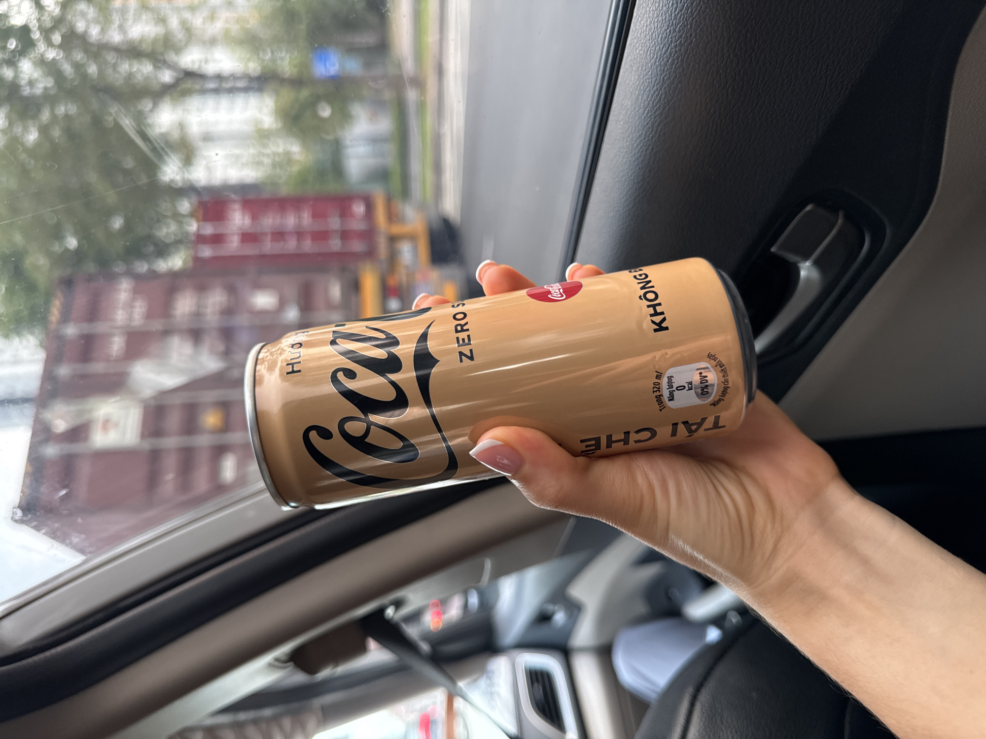 A hand holds a tan "Coca-Cola Zero Sugar" can inside a vehicle. The label is partly in Vietnamese. The background shows a blurred view of a city street, including stacks of shipping containers, seen through the car window. The interior of the car features black upholstery and dashboard elements. The can reads "TAI CHE" and "KHONG" in Vietnamese.
