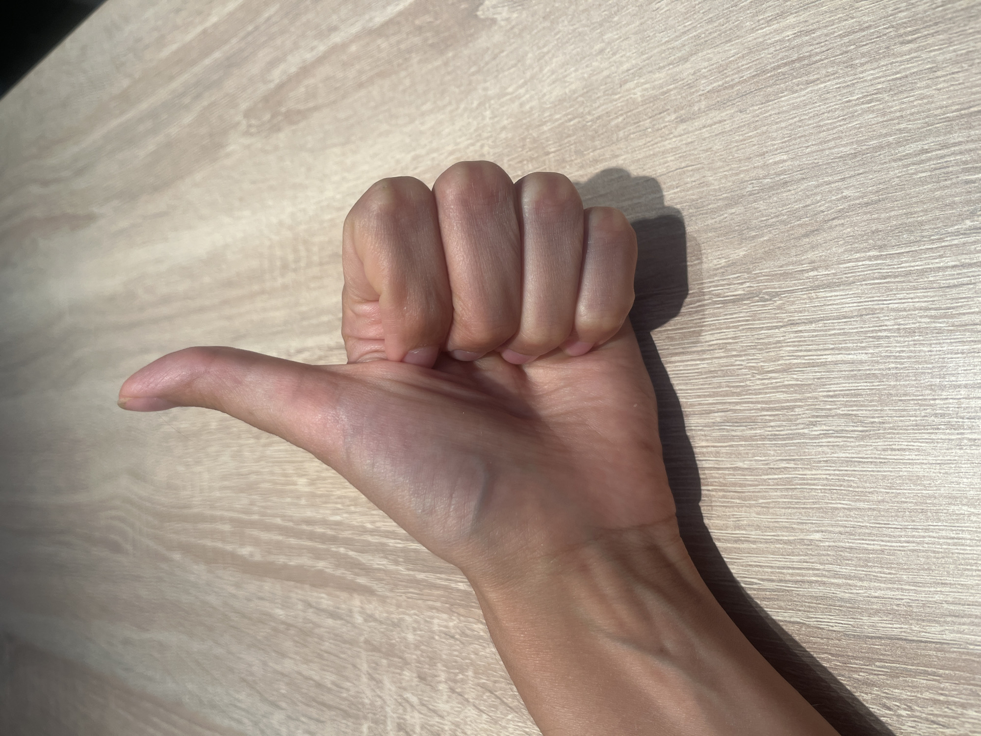 A close-up, eye-level shot shows a person's left hand giving a thumbs-up gesture on a light-colored wooden surface. The hand is positioned in the lower left quadrant of the frame, with the thumb pointing upwards. The fingers are curled into a fist, and the palm is facing the viewer. The wooden surface has a light grain pattern. The lighting creates a shadow of the hand on the surface.