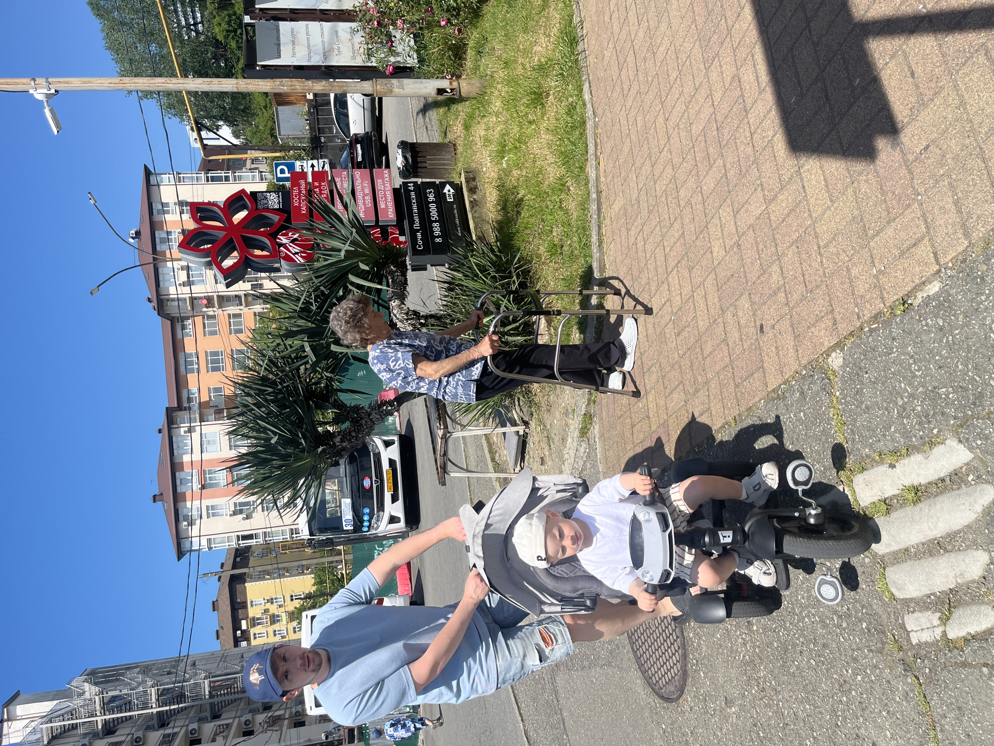 The image captures a bright, sunny daytime scene on an urban sidewalk in Sochi, Russia, specifically near Poltavskaya 44. The sky is clear and blue, suggesting pleasant weather.
In the foreground, a man in a light blue t-shirt, ripped jean shorts, and a blue baseball cap pushes a young child seated in a grey and white tricycle that resembles a toy car. The child, wearing a white shirt and cap, looks towards the camera. Further right, an elderly woman with short, curly grey hair sits on a folding chair, resting beside a silver walker. She is dressed in a patterned blue and white top and dark pants.
The background features several multi-story buildings, some with balconies and air conditioning units. Palm trees and other green foliage line the street and sidewalk. Various black signs with white text advertise local businesses and services. A prominent red and black floral-shaped sign, possibly a logo or decoration, stands out among the greenery. A white van is partially visible behind the elderly woman, with a license plate "AK 214 23".
Visible text on the signs indicates businesses such as a "КАПСУЛЬНЫЙ ХОСТЕЛ" (Capsule Hostel) offering "ИНДИВИДУАЛЬНО USB, Wi-Fi" (Individual USB, Wi-Fi) and "МЕСТО ДЛЯ ХРАНЕНИЯ БАГАЖА" (Luggage Storage). Another sign lists services like "Ногтевой сервис" (Nail service), "Наращивание ресниц" (Eyelash extensions), "Парикмахер-стилист" (Hairdresser-stylist), "Турбо Солярий" (Turbo Solarium), and "Шугаринг Вакс" (Sugaring Wax). The address "Сочи, Полтавская 44" and a phone number "8 988 5000 963" are also displayed, along with a QR code. The ground is paved with tan bricks on the sidewalk and asphalt on the street, with a manhole cover visible near the tricycle. Strong shadows indicate a high sun angle.