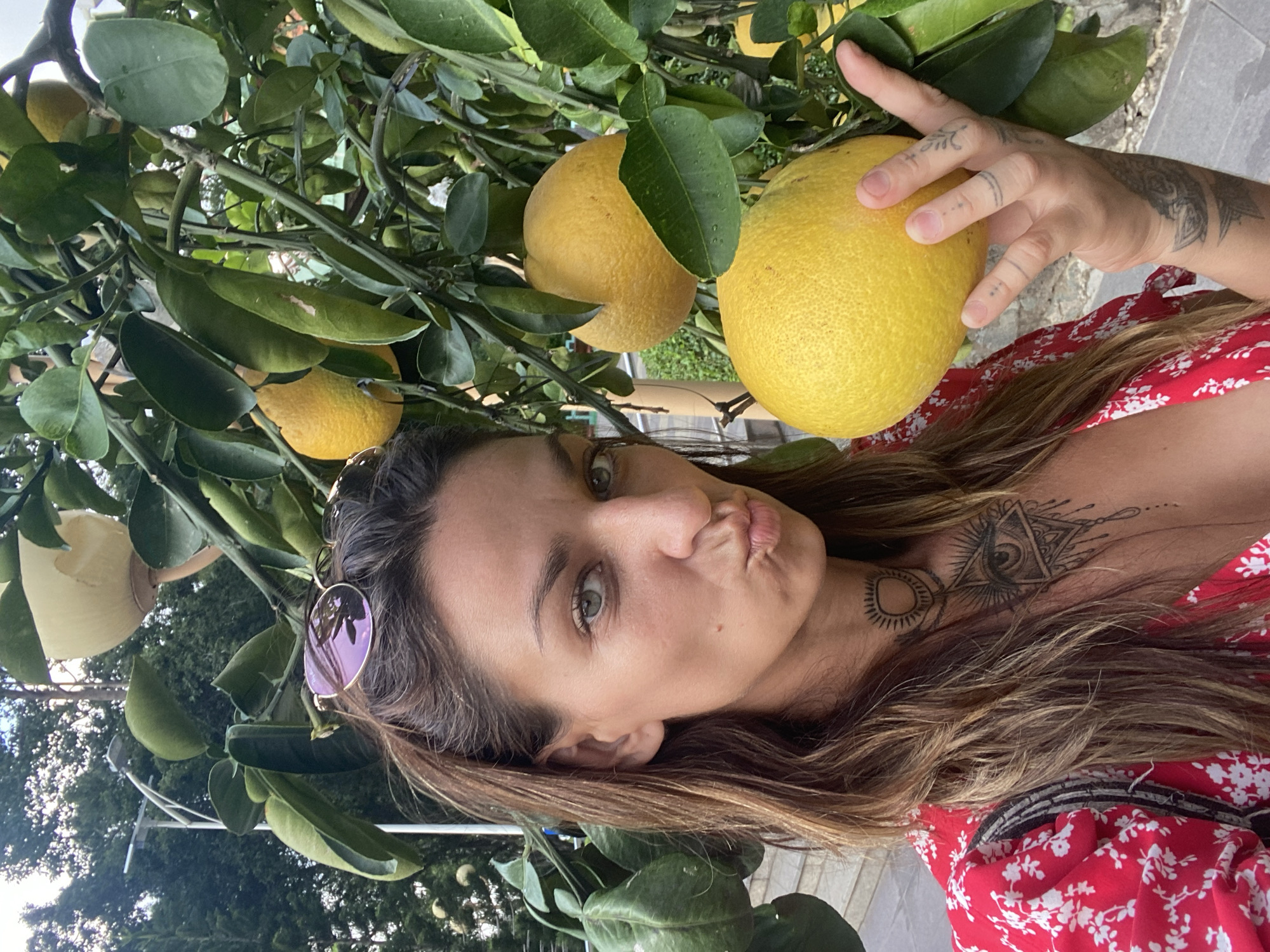A woman with light brown hair and tattoos poses outdoors with a pomelo tree. She is wearing a red and white floral dress and has sunglasses perched on her head. She is puckering her lips. Her left hand holds a large pomelo fruit. Other pomelos and leafy branches are visible behind her and to the left. The background shows a garden environment with hints of architecture. The image was taken in a landscape orientation. There is no evidence suggesting it was taken in Ho Chi Minh City, Vietnam.