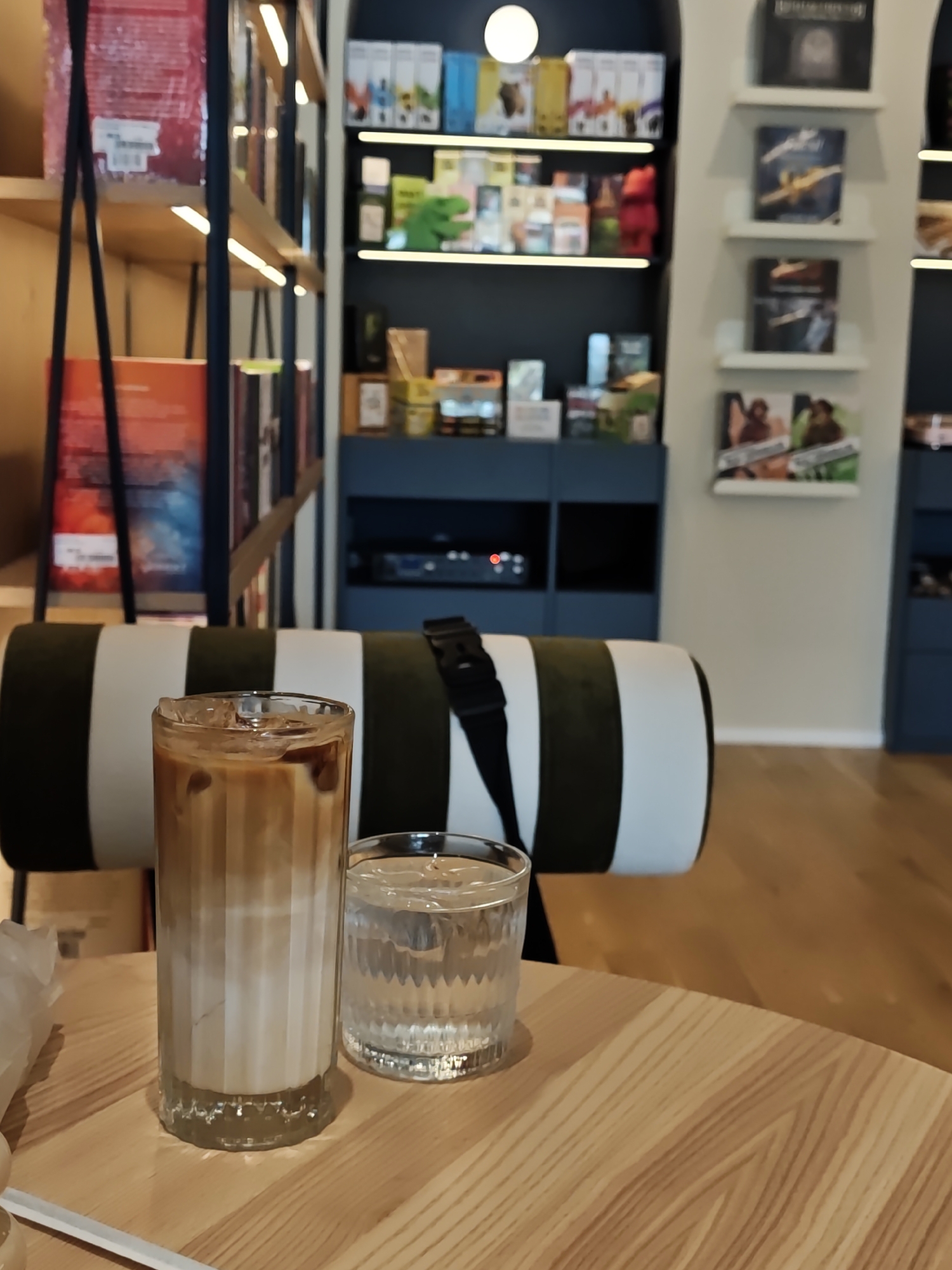 The image shows a close-up view of two glasses on a wooden table, with a striped cushion and shelves of books and other items in the background. The glass in the foreground is a tall, fluted glass filled with an iced coffee drink that has layers of milk and coffee. The glass next to it is a shorter, rippled glass filled with ice and water. The table is made of light-colored wood with a visible grain. In the background, a cushion with black and white stripes is visible, possibly part of a seating area. Behind the cushion, shelves are filled with books, boxes, and decorative items. The shelves are illuminated by overhead lights. The overall setting appears to be a cafe or a library. The image does not provide any specific visual cues to determine the exact location in Kyiv, Ukraine.