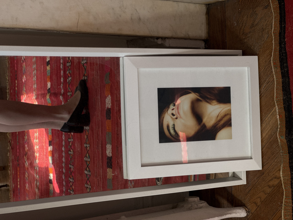 This scene is located in a room within New York City, United States of America. A large, white-framed, rectangular mirror stands upright, reflecting the lower legs and feet of one individual. The individual is wearing dark, closed-toe shoes with block heels. The floor is covered by a red, patterned rug featuring geometric designs in various colors, including orange, light blue, dark blue, white, and black stripes. A dark wooden floor is visible at the bottom of the frame. Leaning against the mirror are two white-framed objects. The most prominent is a photograph depicting a close-up portrait of a person, likely female, with light brown hair, looking upwards with eyes closed or partially closed. A horizontal streak of light reflects across the center of this photograph. A second, similar white-framed object is partially visible behind and to the left of the main framed photograph. On the far left, a portion of a light-colored furniture piece is visible. On the far right, a textured, light-colored wall or column is present. Natural light, possibly sunlight, illuminates parts of the reflected rug with a warm, reddish glow.