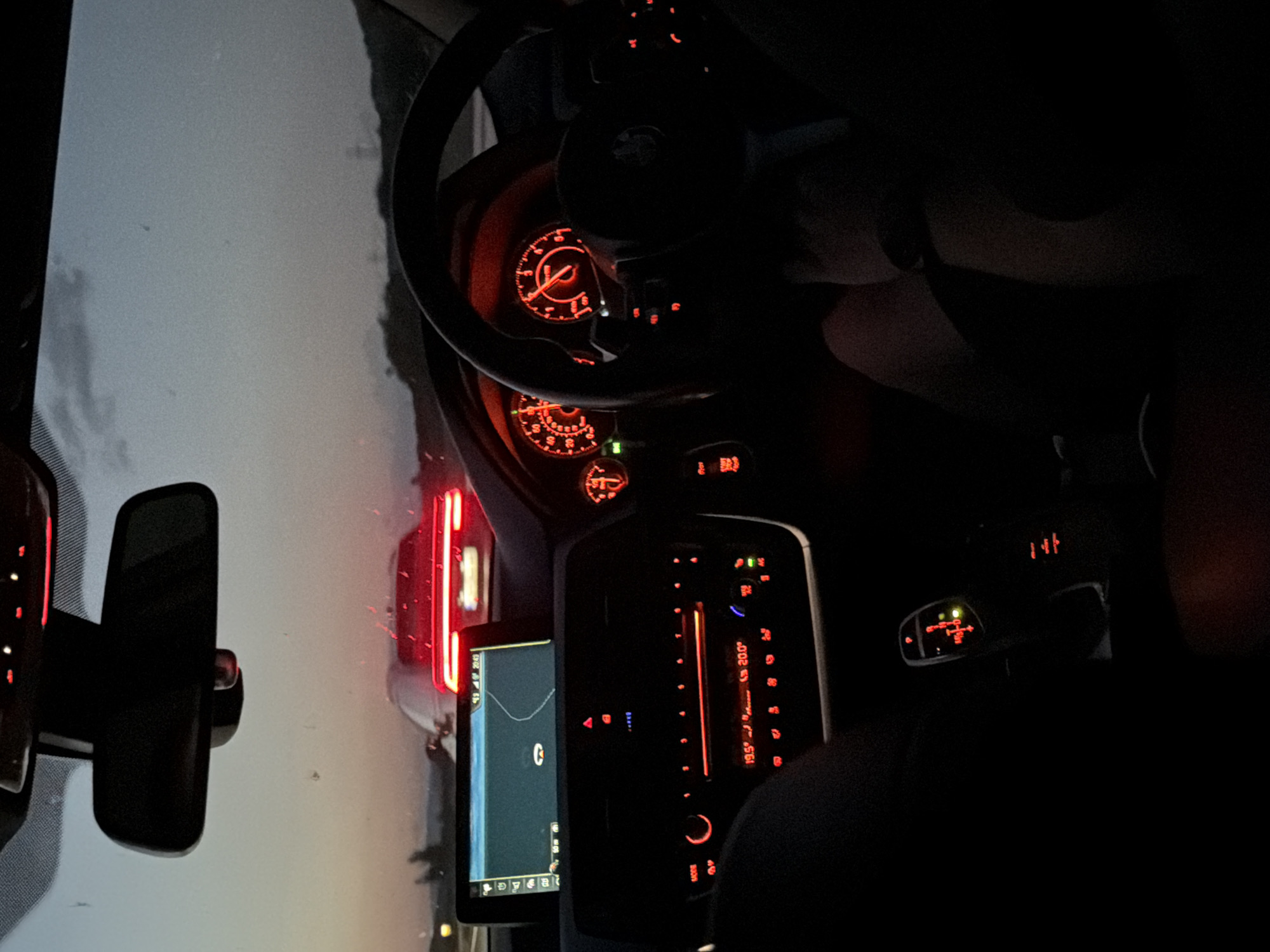 The image depicts an interior perspective from a BMW vehicle at dusk or night, looking forward through the windshield. The vehicle's dashboard, instrument cluster, and center console controls are illuminated in red. A driver's hand, wearing a watch, rests on the black multi-function steering wheel, which prominently displays a BMW emblem. A passenger's leg and arm are partially visible in the bottom right.
The instrument cluster features two main analog gauges, illuminated in red, alongside a smaller gauge to the left. A central infotainment screen displays a navigation map with a highlighted route, showing a battery charge of "93%" and the time "20:49". Below the screen, the climate control and audio system display temperature settings of "19.5°" and "20.0°". The illuminated electronic gear selector indicates "P" and "M/S" options with a green light suggesting an engaged drive mode. The rearview mirror is visible at the top center, reflecting the ambient light.
Outside the vehicle, a red car is visible ahead on what appears to be a highway or multi-lane road. Its distinct, horizontal brake lights are illuminated. The sky above the horizon shows residual twilight, with dark, indistinct landforms or structures in the distance. The license plate of the car ahead is unreadable.
Based on the visual evidence, the car is likely stationary or moving slowly. The metric temperature units and vehicle type are consistent with European use. However, there are no specific visual cues or identifiers within the image (such as recognizable landmarks, legible road signs, or discernible license plate details) to forensically confirm the precise location as Mursalevo, Bulgaria.
