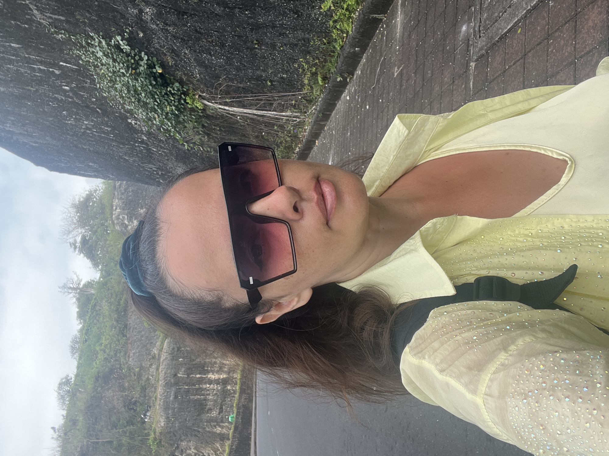 A woman stands on a paved walkway next to a road with a rocky, vegetated cliff face behind her. She is wearing a pale yellow, collared, sheer blouse adorned with iridescent sequins over a matching camisole. She has dark hair pulled back in a ponytail with a dark teal scrunchie and is wearing large, square sunglasses with pink-tinted lenses. A dark backpack is visible on her shoulder. The environment suggests Kuta Selatan, Indonesia, with its characteristic road infrastructure and rocky outcrops. The sky is overcast.