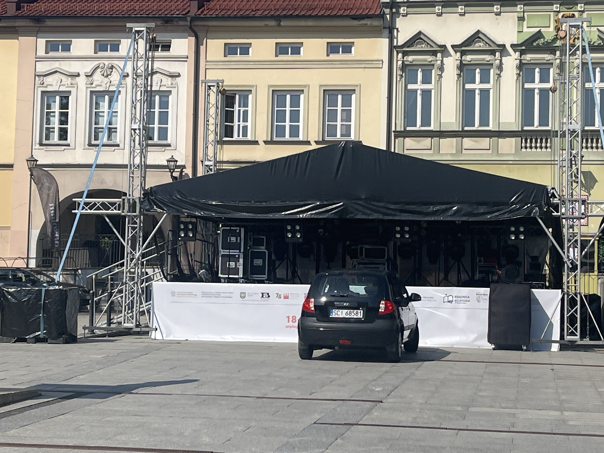 The image depicts an outdoor stage setup in a town square or plaza. The stage is covered with a large black tarp, and behind it, sound equipment and lighting rigs are visible. In front of the stage, a white banner with logos and text is displayed. A black hatchback car is parked in front of the banner, facing away from the viewer.
The setting appears to be a historic European town, judging by the architecture of the buildings in the background, which feature ornate windows and classical facades. The buildings are painted in various light colors, including yellow and cream, and have red tile roofs. The ground is paved with grey concrete slabs.
The time of day appears to be daytime, with bright sunlight casting shadows on the ground, suggesting a sunny day. The overall atmosphere seems to be neutral, possibly awaiting an event.
Visible text on the banner includes Polish words such as "Biblioteka" and "Kultura," indicating the event may be related to library or cultural activities. There are also several logos on the banner, one of which is a book with an open page. The car's license plate is visible and reads "SCI 68581".