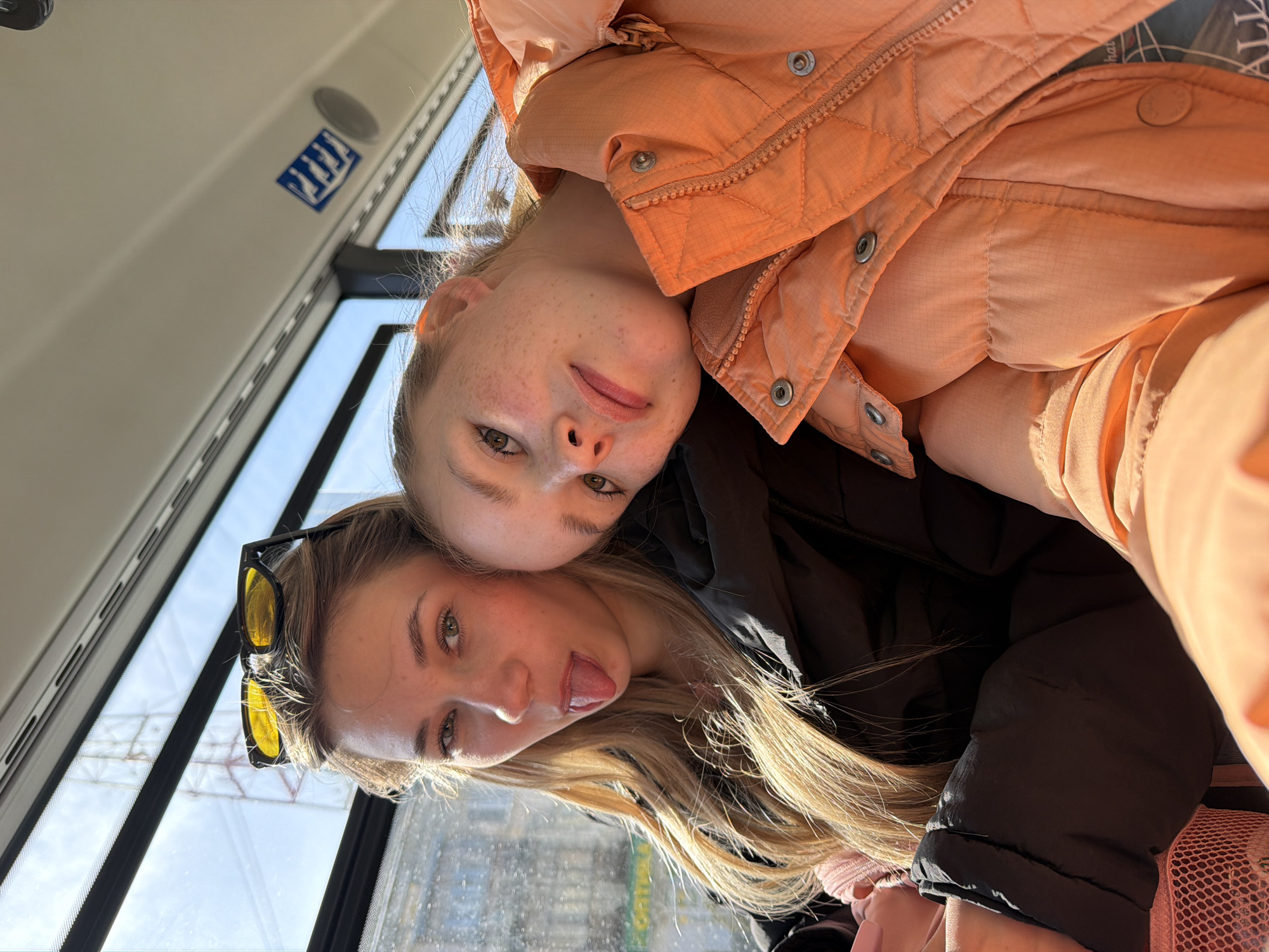 Two Caucasian females pose closely for a selfie inside a public transportation vehicle. The female on top has blonde hair with freckles and is wearing an orange puffer jacket. The female underneath has long blonde hair and is wearing sunglasses on top of her head, and is sticking her tongue out. Through the windows, glimpses of the urban environment of Berezniki, Russia are visible. The window has some water droplets visible. A sticker with symbols is visible at the top of the image near the window.