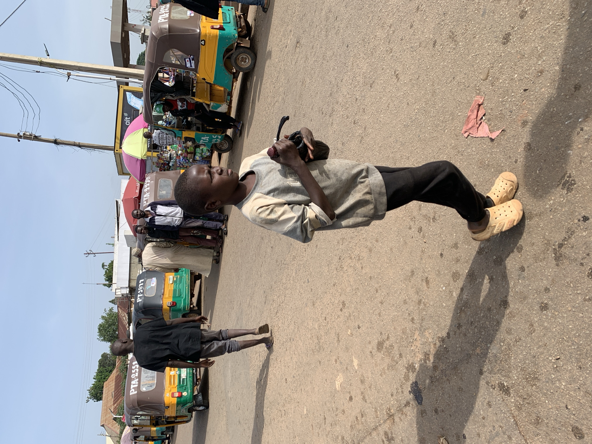 The image depicts a street scene in Gura Top, Nigeria, with multiple auto-rickshaws (keke napep) parked on a dirt road. In the foreground, a young boy wearing a light-colored long-sleeved shirt and black pants stands facing left, holding a black bag and a red object. He is wearing light yellow clogs. Behind him, an adult male in a black t-shirt and gray pants walks away from the camera, also wearing sandals. Other individuals and parked auto-rickshaws are visible in the background, along with market stalls and utility poles. The sky is clear and blue, suggesting a daytime setting. The ground is dusty with scattered debris.