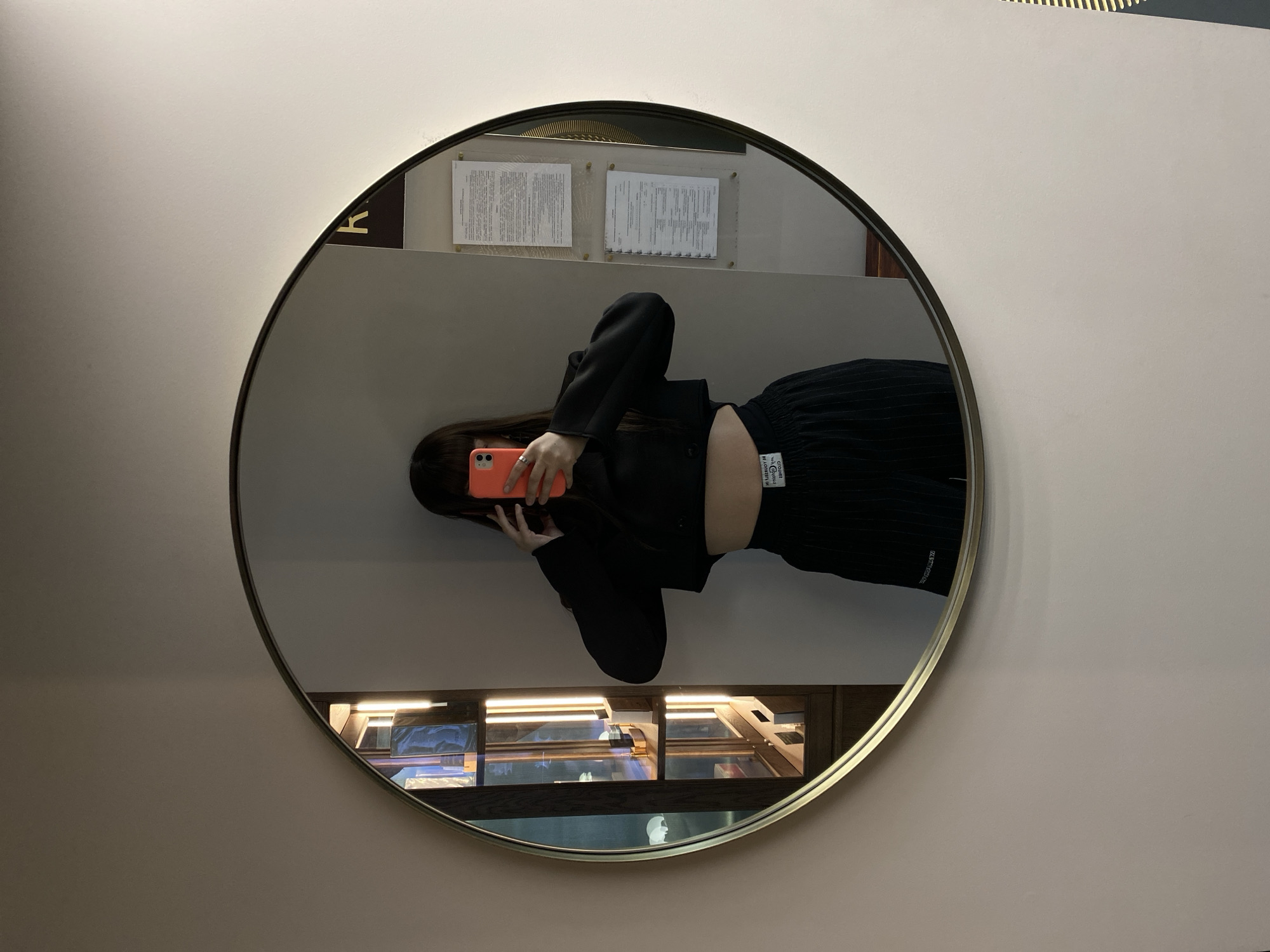 The media file presents a mirror selfie taken by a person in an indoor setting. The image is oriented 90 degrees clockwise, showing the subject lying horizontally. A circular mirror, framed with a thin gold-colored rim, is mounted on a plain, light beige or off-white wall.
The subject is a person, likely female, with long dark hair, dressed in a black long-sleeved cropped top or jacket and black pleated trousers. Their face is obscured by a vibrant orange smartphone held in their hands, used to capture the reflection. A silver ring is visible on one of their fingers.
The reflection in the mirror reveals elements of the surrounding environment. In the upper part of the reflection, above the person's head, two framed documents with illegible text are visible on a wall. A partial golden letter "K" is also discernible near these documents, possibly part of a sign or logo. Below the person's waist in the reflection, there is a wooden display case or shelving unit, illuminated by small overhead lights, showcasing various items, which appear to be books or small artifacts.
Visible text on the waistband of the person's trousers clearly reads "COUTURE" and "AUTHENTIC" in white letters on a black label. The interior setting, with its display case and informational documents, suggests a public space such as a gallery, museum, or shop. The time of day is not evident, as the scene relies on indoor lighting. The location context is Votkinsk, Russia.