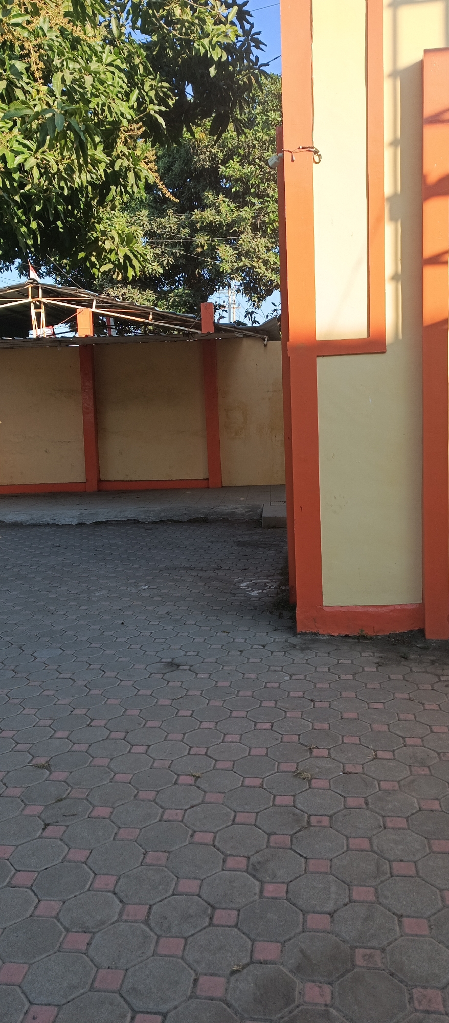 The image depicts an outdoor scene in Arjasa, Indonesia, featuring a paved area in the foreground and a wall with a gate-like structure on the right. The ground is covered with octagonal paving stones, alternating with smaller square pink stones, forming a patterned surface. A wall, painted in a pale yellow with orange trim, runs horizontally across the middle ground. To the right, a pillar of the same yellow and orange design stands prominently, with a small, metallic object, possibly a lock or latch, attached near its top. Beyond the wall, lush green foliage from trees is visible against a clear blue sky. A portion of a rooftop structure, with corrugated metal sheeting, is situated behind the wall.