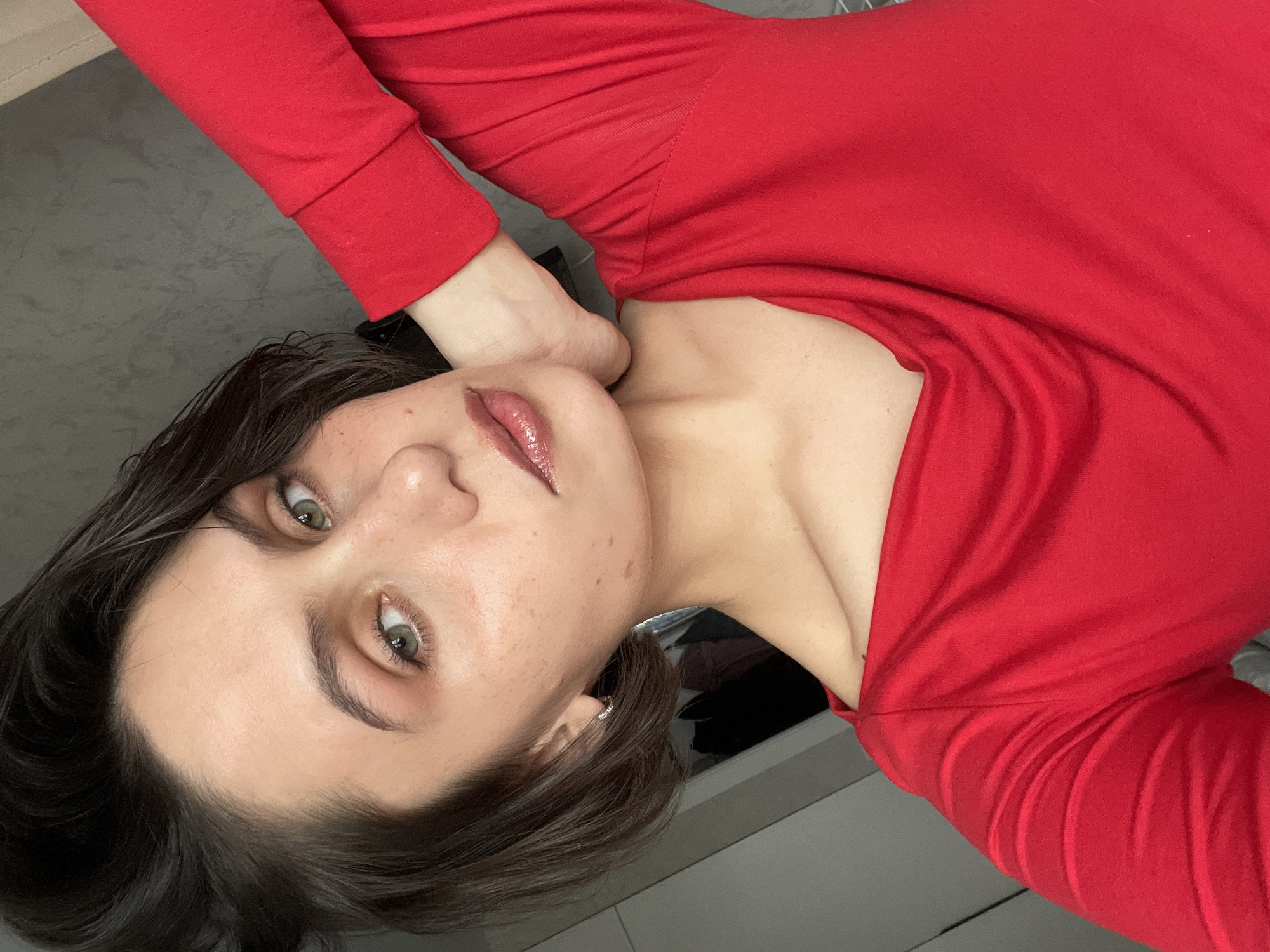 A woman with fair skin, short brown hair, and hazel eyes is posing for a close-up portrait. She is wearing a red, long-sleeved garment. Her hand is placed near her neck. The background appears to be a neutral gray surface. There is no evidence in this image to indicate it was taken in Izhevsk, Russia.