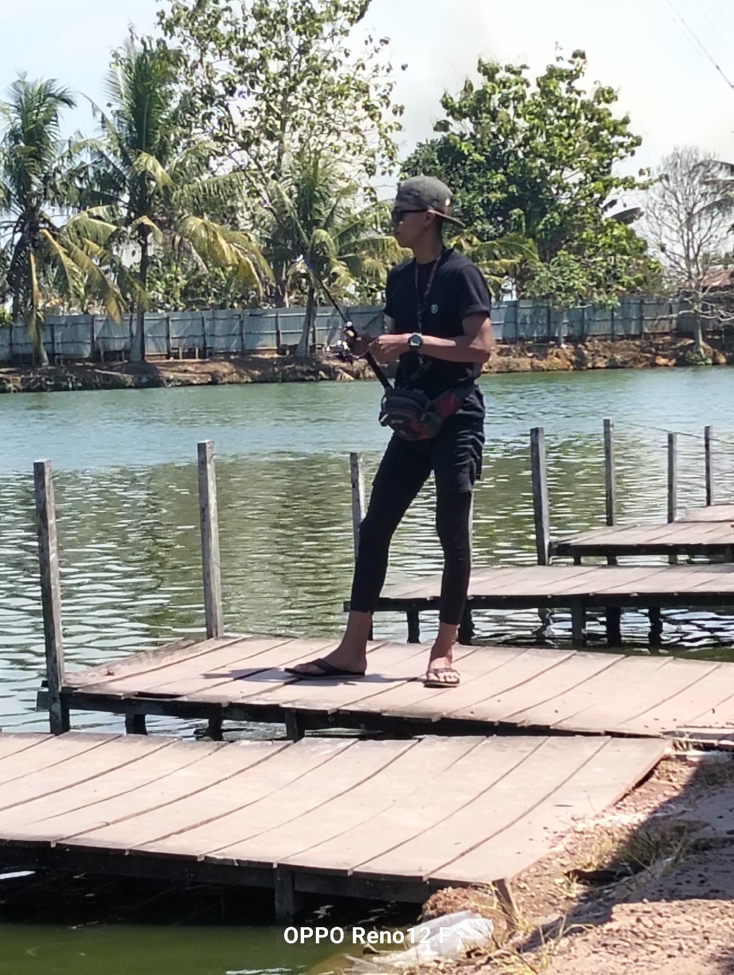A man stands on a wooden pier extending into a body of water. He is wearing a dark t-shirt, black pants, a baseball cap, sunglasses, and flip-flops. He holds a fishing rod and has a fanny pack around his waist. The water is calm and greenish. Palm trees and other vegetation are visible in the background, along with a fence. The scene appears to be located in the city of Cindai Alus, Indonesia, as indicated by the text overlay at the bottom of the image. The lighting suggests it is daytime with clear weather.