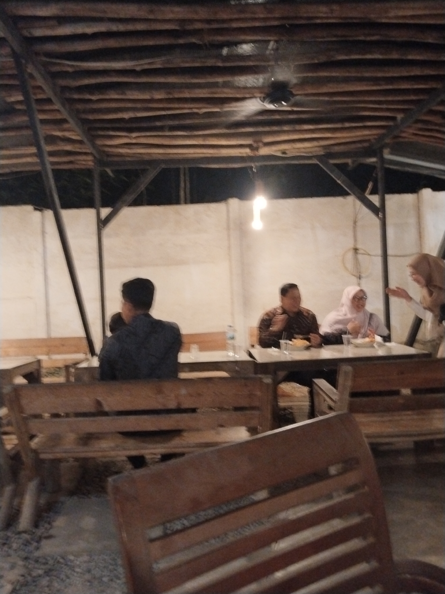 In an outdoor dining area with a wooden plank ceiling and metal supports, a ceiling fan is visible. Three individuals are seated at a table in the background. A man wearing a patterned shirt is on the left, facing a woman in a light pink hijab and glasses, who is on the right. Another person, wearing a light brown hijab, is standing on the far right, gesturing with their hands. They appear to be engaged in conversation. In the foreground, wooden benches are arranged, and a wooden chair is prominently in view. The lighting is dim, suggesting it is evening. The ground appears to be gravel or dirt. The scene is likely at a casual eatery or restaurant in Serang, Indonesia, given the attire of the people and the rustic outdoor ambiance.