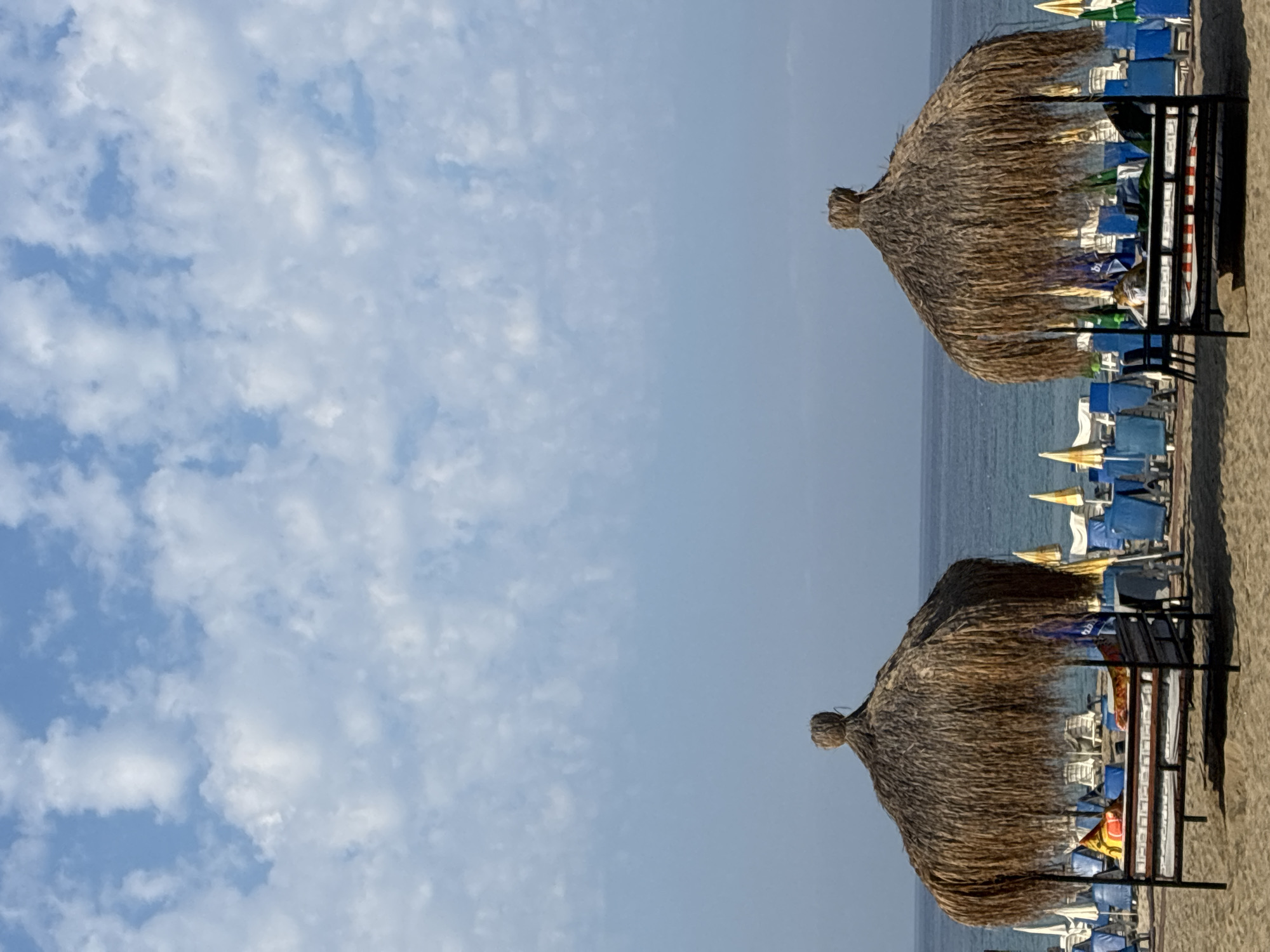 This is a vertical, eye-level, medium shot of a sandy beach on a sunny day. The sky is blue with scattered white clouds. The bottom half of the image is dominated by the beach, with two thatched-roof cabanas in the foreground. Each cabana is a raised platform with a thatched roof and appears to have cushions or bedding on it, suggesting it's for lounging. In the background, there are rows of blue lounge chairs and a few yellow and white umbrellas, indicating a resort or public beach setup. The sea is visible behind the beach, with a calm surface and a clear horizon. The overall impression is one of a relaxing beach scene in Alanya, Turkey, a popular tourist destination known for its beaches.