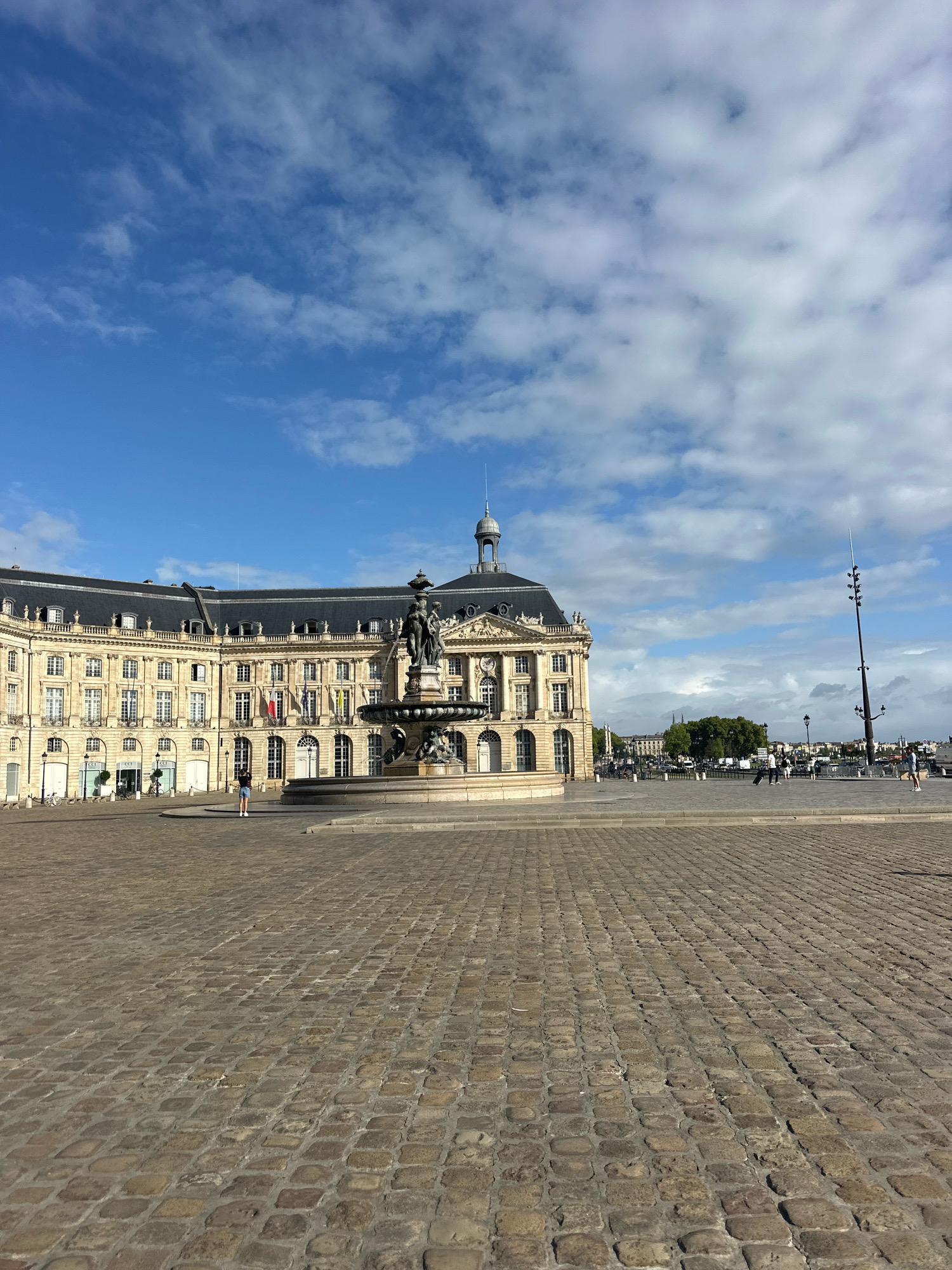 The image depicts the Place de la Bourse in Bordeaux, France, on a sunny day with scattered clouds. A large, ornate building with a curved facade, characteristic of 18th-century architecture, dominates the background. The building features numerous windows, classical columns, and a dark, mansard roof topped with a cupola. In the foreground, a grand fountain with sculptures and multiple tiers is centrally positioned. The ground is paved with cobblestones, creating a textured surface that extends across the open square. Several people are visible in the middle ground, walking and standing, including one person in the foreground taking a photograph. A lone figure stands near the fountain, facing the building. The sky is blue with dramatic white clouds. The scene conveys a sense of historical grandeur and public space activity.