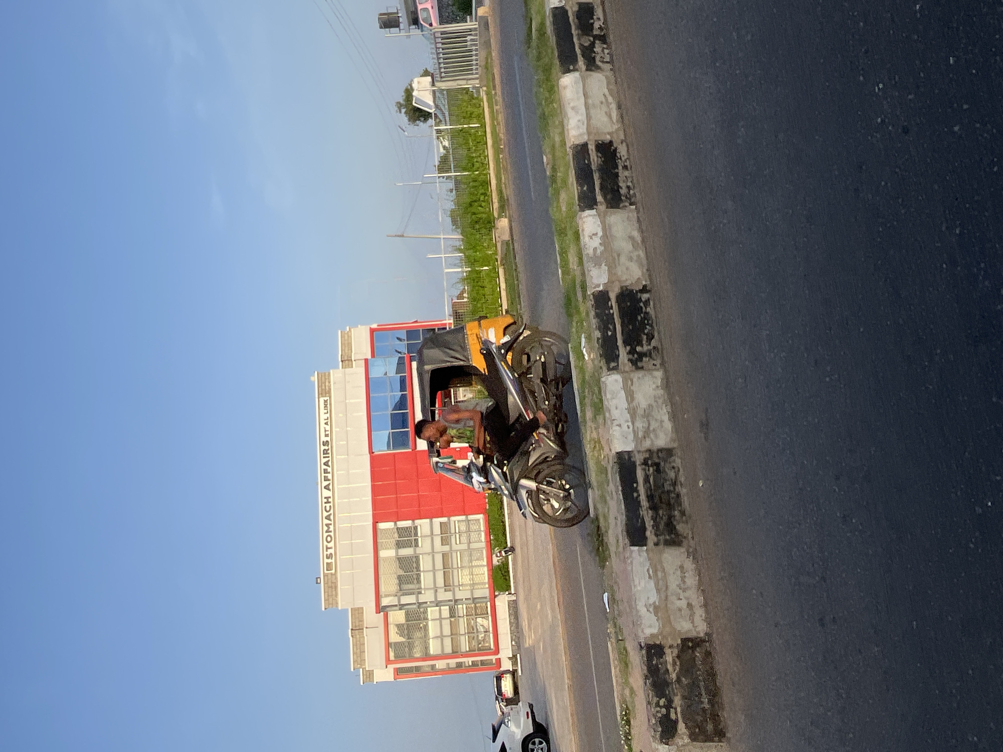 A man rides a motorcycle with a passenger and a sidecar in an urban setting. The motorcycle is silver and black, and the passenger is wearing a green shirt and dark pants. The sidecar is orange and black. The man is looking forward, and the passenger is looking to the right. In the background, there is a modern building with red and white accents. The building has large windows and a sign that says "STOMACH AFFAIRS IT AL LINK". There is a street in the foreground with a concrete barrier separating it from a patch of grass. The sky is clear and blue.