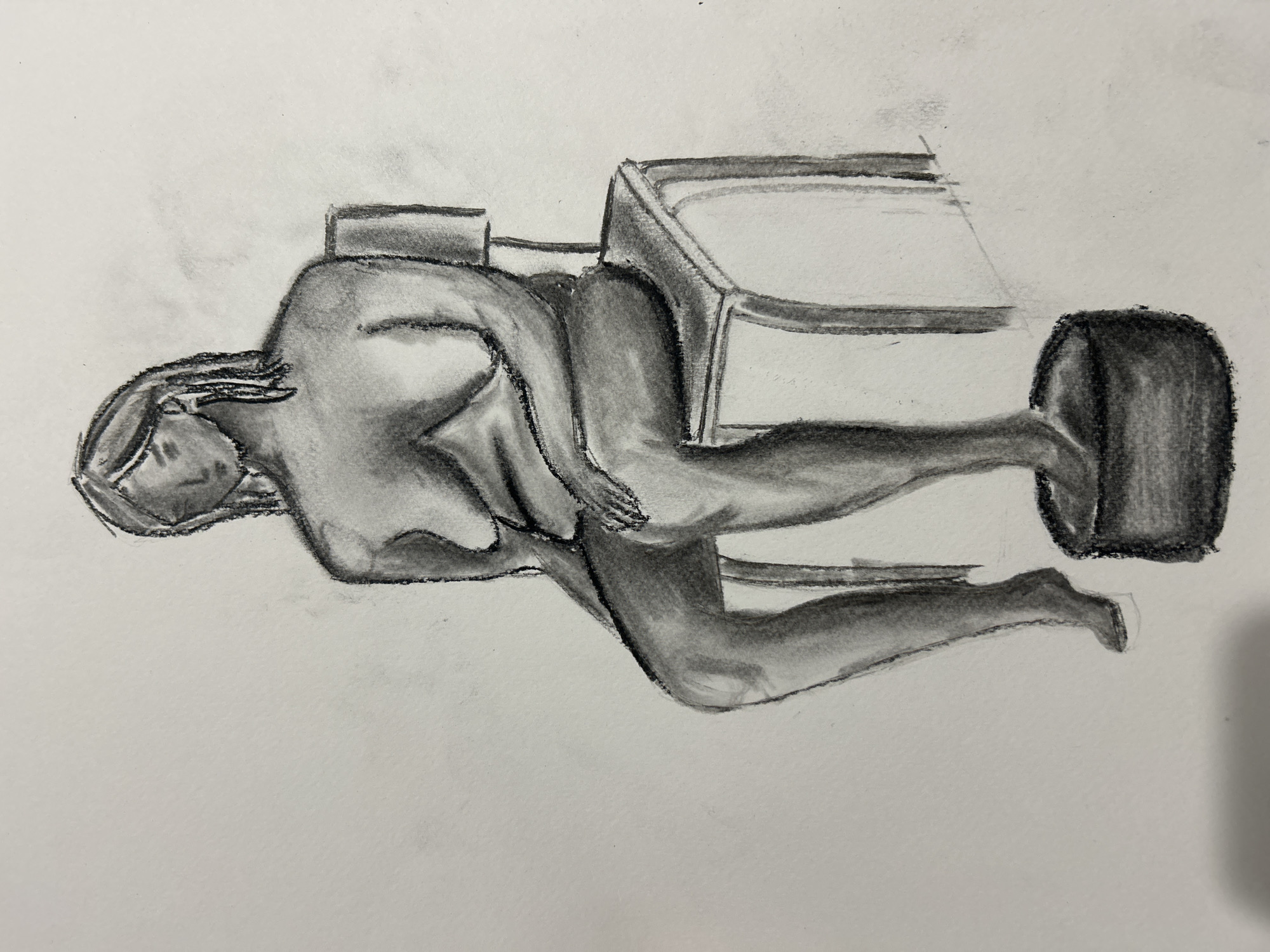 A charcoal drawing depicts a person, possibly a nude female figure, bent over and carrying a box-like structure on their back. The box resembles a small, crude rendering of a piece of furniture. The figure is facing left and seems to be burdened by the weight of the object. The drawing is rendered in grayscale, with varying shades of gray and black to create depth and form. There is no indication that the scene is located in Sofia, Bulgaria.