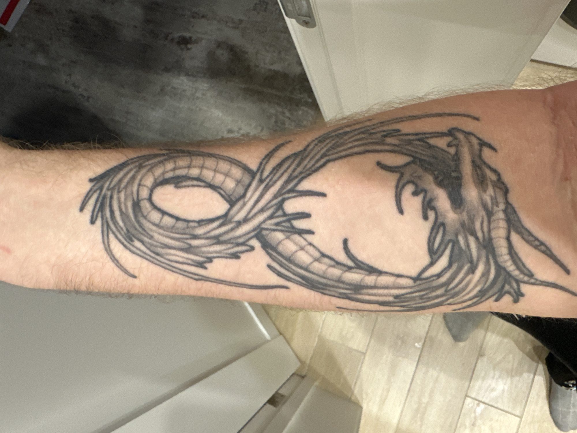 A tattoo of a dragon in the shape of an infinity symbol on a person's forearm is depicted. The forearm is pale skin with light-colored hair. The tattoo is black ink. The background includes a gray floor or surface and a white object that appears to be furniture. The location is not discernible as Oryol, Russia, from the image alone.