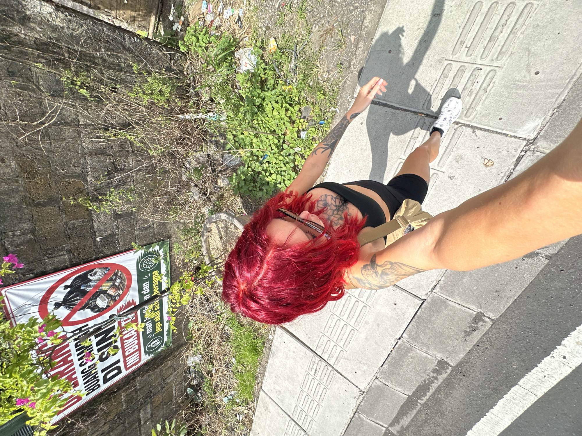 A high-angle shot depicts a person with bright red hair, wearing a black top and black cycling shorts, standing on a concrete sidewalk. The person is fair-skinned, with tattoos visible on their arms and chest. Their right arm is outstretched towards the camera, indicating a selfie. On the left side of the frame, there is a stone wall covered with greenery and debris, suggesting a somewhat neglected urban environment. Attached to the wall is a sign prohibiting the dumping of garbage, with the words "DILARANG BUANG SAMPAH DISINI" visible, indicating the location is in Indonesia. The sidewalk includes tactile paving intended for pedestrians with visual impairments. The angle and lack of other surrounding elements suggest an urban area. The location is North Kuta, Indonesia, confirmed by the sign on the wall.