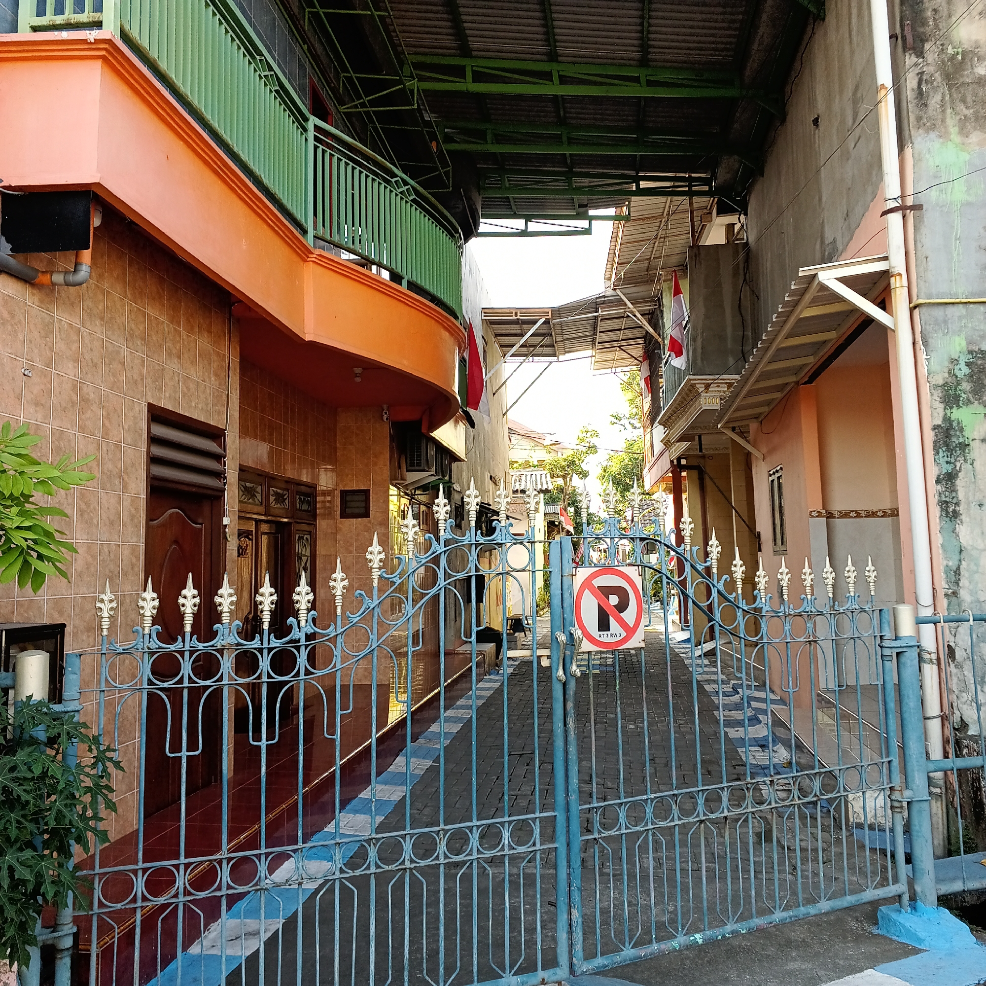 The image depicts a street scene in Wedoro, Indonesia, characterized by a narrow alleyway flanked by buildings. In the foreground, a blue metal gate with ornate finials stands closed, blocking access to the alley. Attached to the gate is a circular red and white sign with a black letter "P" crossed out, indicating "no parking."
The buildings on either side of the alley exhibit a variety of architectural styles and materials. The building on the left features a tiled facade and an orange-colored upper story with a green metal railing. The building on the right has a rough concrete exterior and a pale pink lower section. Overhanging roofs and awnings made of corrugated metal provide shade over the alleyway.
A red and white Indonesian flag is visible hanging from the building on the right, suggesting a national or celebratory context. The ground of the alleyway is paved with dark stones and has a checkered pattern of white and blue stripes. A lush green plant with large leaves grows on the left side of the frame, partially obscuring the tiled wall and a dark wooden door. The lighting suggests it is daytime, with sunlight casting shadows along the alley. There are no people visible in the image.