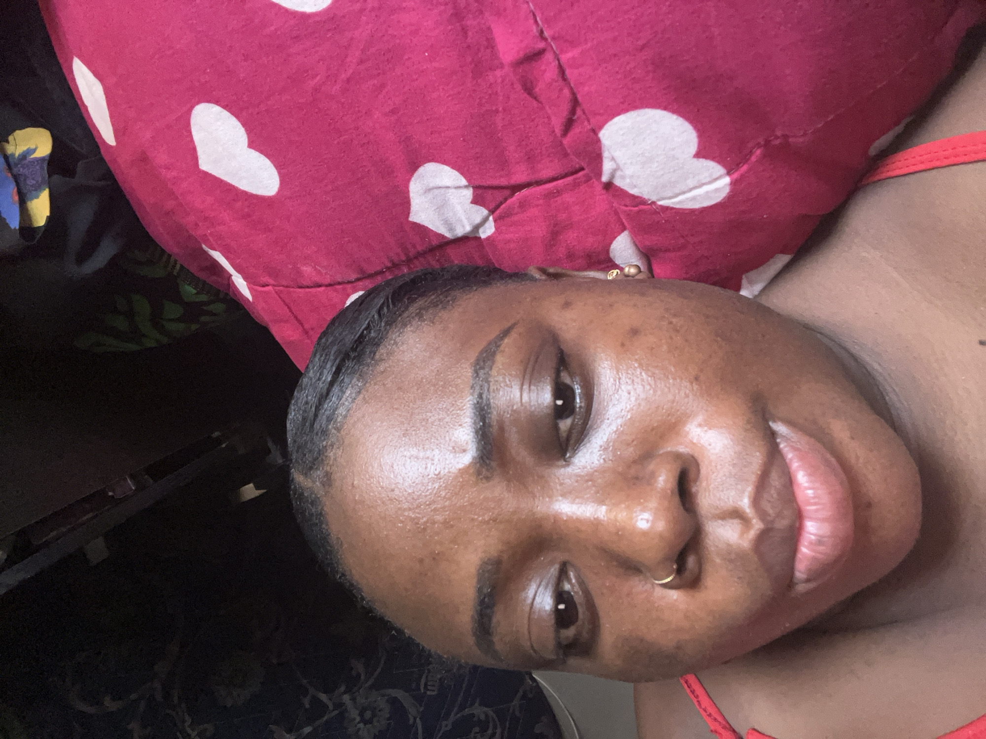 A close-up photograph depicts a dark-skinned person with a gold nose ring and gold earrings lying down. The person is resting their head on a red pillow with white heart patterns. The background is dark, revealing parts of patterned fabric and a metallic shelf. The location cannot be determined from the image, nor can any details regarding the city of Abuja, Nigeria.