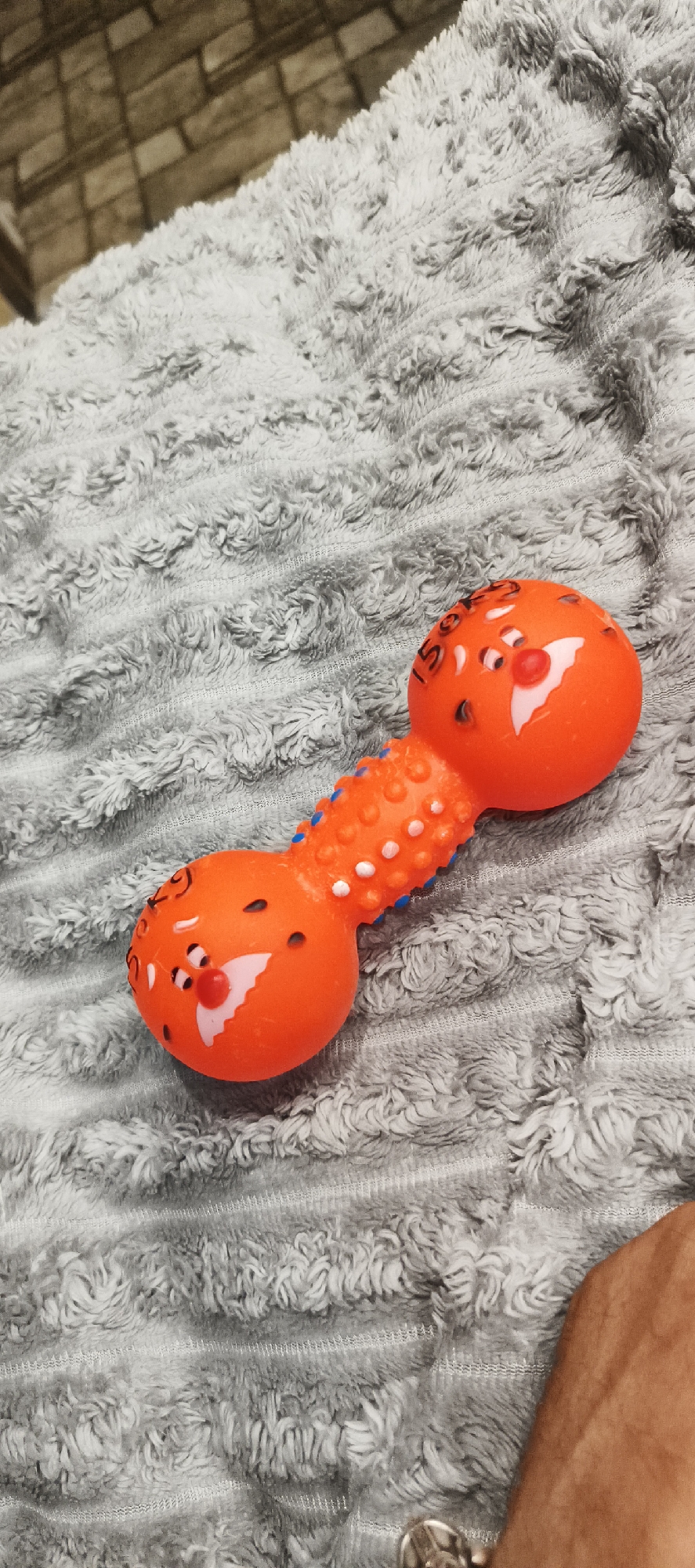 An orange dog toy, shaped like a dumbbell with round ends, lies on a textured gray blanket. The toy features painted cartoonish faces on each end with red noses, black eyes, and white accents. The center of the toy has a textured, ridged section with small blue and white dots. The background is blurred, showing a brick pattern. There is a portion of a human arm visible in the lower right corner. The location is Liubashivka, Ukraine.