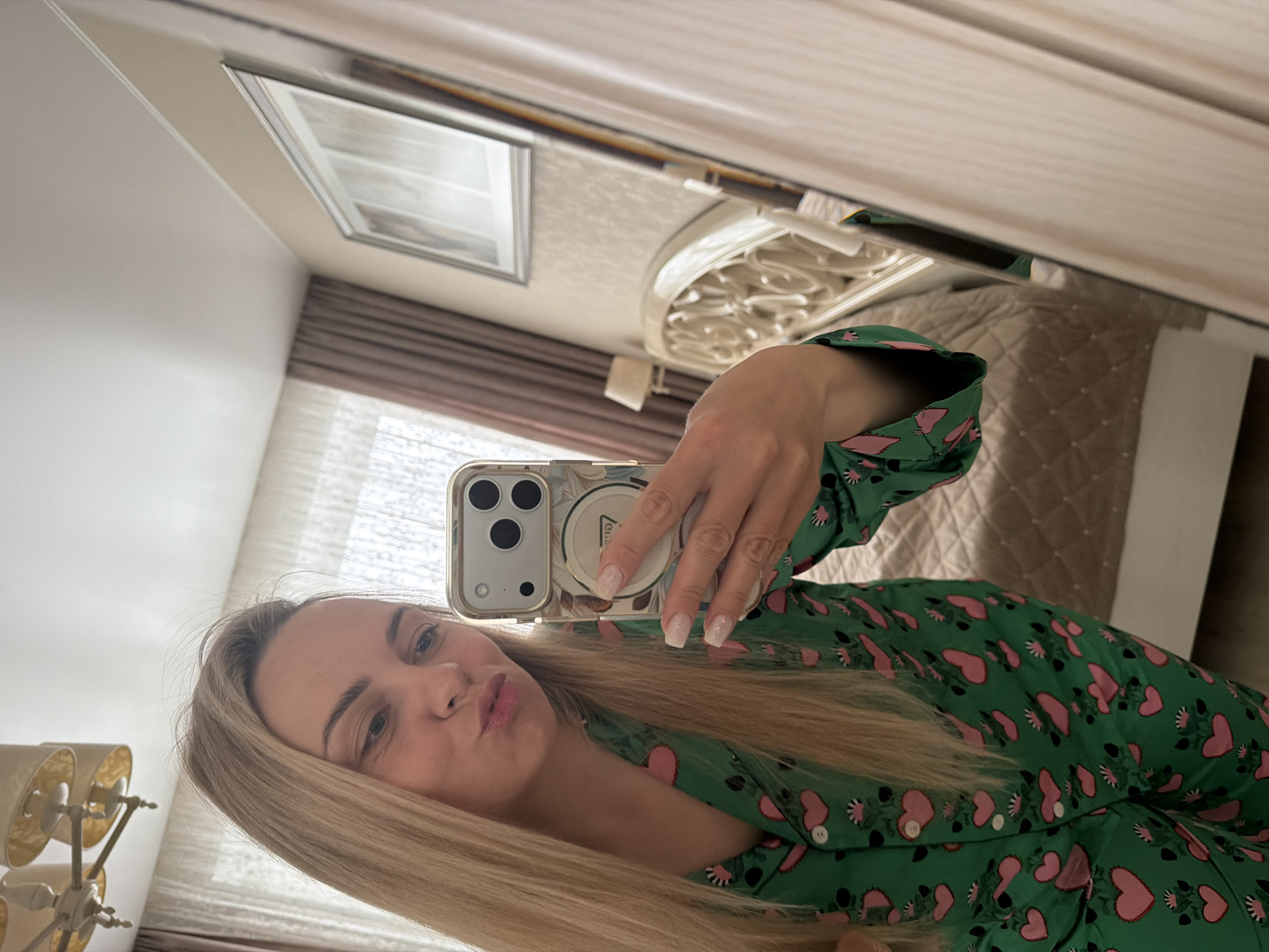 This media file is a mirror selfie featuring a fair-skinned woman with long, straight blonde hair and light eyebrows. She is wearing a green long-sleeved top or pajama set patterned with pink hearts and small green and white floral elements. Her right hand holds a light-colored smartphone, likely an iPhone, encased in a decorative case that appears to have a subtle floral or abstract design in beige and brown tones. Her fingernails are manicured with a light, possibly glittery, polish. She poses with a slight pout, looking towards her reflection.
The setting is an indoor room, visible through the mirror as the background. It appears to be a bedroom with a bed featuring a light-colored, quilted bedspread and an ornate white or light-colored headboard with intricate scrollwork. Light-colored curtains are visible, suggesting a window. To the lower left of her face, a wall-mounted lamp with two cream-colored fabric shades is present. A framed picture hangs on the wall above the bed. The room is brightly lit, indicating daytime. The image itself is rotated approximately 90 degrees counter-clockwise. The location context for this image is Votkinsk, Russia. An illegible brand logo is present on the back of the phone case.