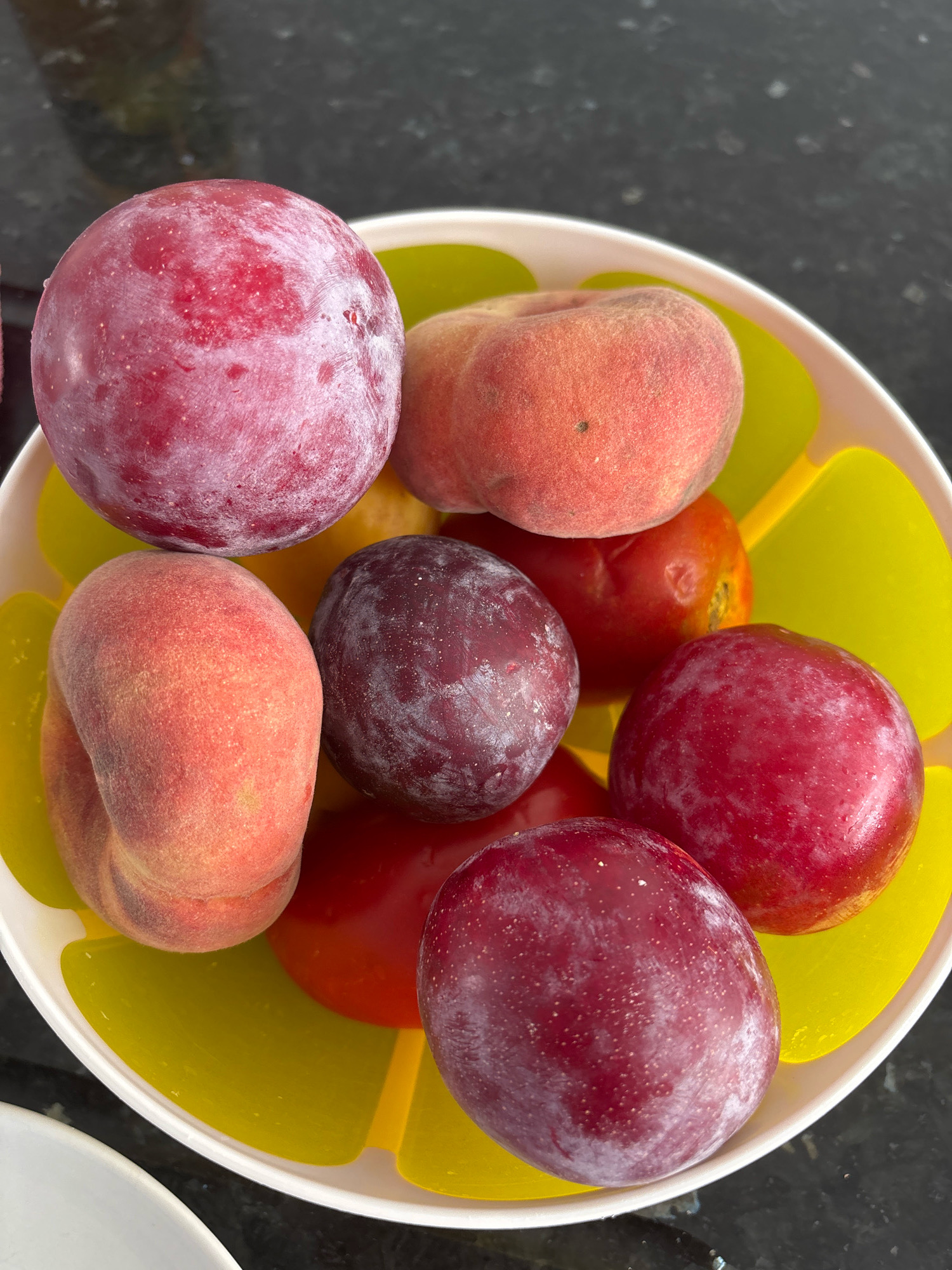 A close-up, overhead shot captures a vibrant medley of fruits in a white and yellow bowl, set against a dark, speckled countertop. The bowl itself has a distinctive yellow insert with petal-like shapes.
The fruits appear to be a mix of plums and peaches, with one object in the background that could be a tomato or another type of plum.
Specifically, there are four plums visible. One large, deep red plum dominates the upper left of the frame, with a slight dusting of white bloom. To its right, a plump, fuzzy peach displays a gradient of pink and orange hues. Below the large plum, another peach, more intensely orange and red, lies on its side. To its right, a darker, almost purple plum is nestled. In the center, a round, red object, possibly a tomato or a dark plum, is partially obscured. Finally, in the lower right corner, another deep red plum with a bloom is clearly visible.
The lighting suggests it is daytime, possibly indoors. The composition is tight, focusing solely on the bowl of fruit and the immediate surrounding countertop. There are no people or any identifiable elements of the city of Málaga, Spain, present in this image.