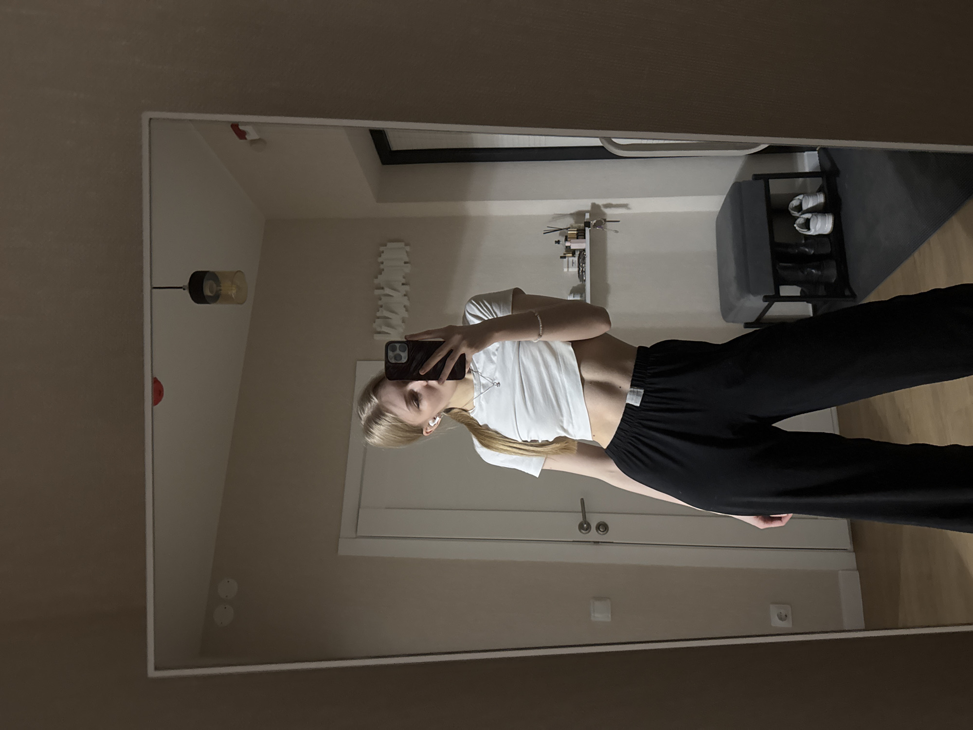 A mirror selfie captures a young person standing indoors, taking their picture in a large mirror with a white frame. The person, likely female, has long blonde hair styled in a braid and wears a white short-sleeved crop top that exposes her midriff, paired with loose-fitting black pants. She has white wireless earbuds in her ears and a silver-colored bracelet on her left wrist. Her face is partially obscured by the dark-cased smartphone she holds up to take the photo.
The background, visible in the mirror's reflection, reveals a modern interior. To the right of the person, a white, multi-tiered shoe rack holds two pairs of shoes, one white and one dark, on a dark floor mat. Above it, a small white shelf or organizer displays various small items. A ceiling light fixture with a glass cylindrical shade and a visible bulb hangs from above. On the left wall, a white, abstract, sculptural decoration is mounted. Behind the person's lower body, a light-colored door with two circular handles and a white electrical outlet is visible. Another white electrical outlet is present on the wall further to the left. A small red object is attached to the top-left inner edge of the mirror frame. The lighting suggests an indoor evening or nighttime setting. The provided location context is Perm, Russia. Text on the white tag of the black pants is not decipherable.