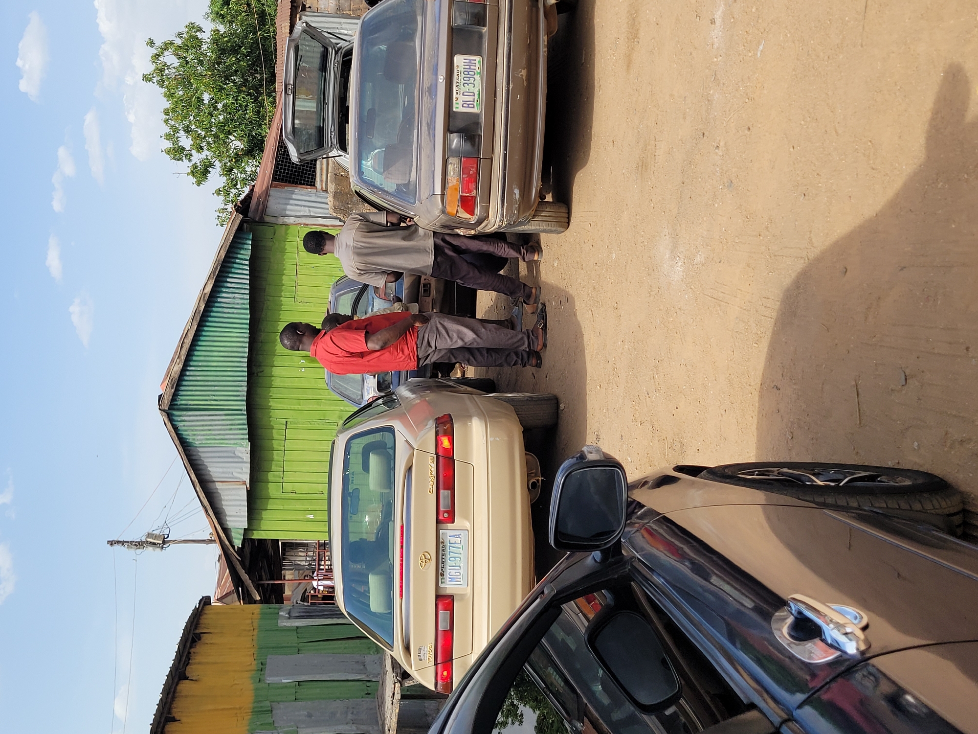 This outdoor scene, likely captured in Jos, Nigeria, depicts an area with several parked cars and simple buildings under a bright, sunny sky. The ground is unpaved and appears dusty.
In the mid-ground, at least three men are present. Two are standing near a gold/tan Toyota Camry sedan, one wearing a red shirt and grey trousers, and the other in a light-colored shirt and dark trousers. Their body language suggests they might be conversing or inspecting the vehicles. Behind them, another man in a light shirt is seen near a dark brown/bronze sedan, which has its trunk open, implying activity such as loading, unloading, or maintenance.
The gold Toyota Camry displays a Nigerian license plate "MGU-977EA" with "FEDERAL" written above it, and car badging for "TOYOTA," "CAMRY," and "16 VALVE." The dark brown sedan also has a Nigerian license plate, "BLD-398HH," with "FEDERAL" above it. Partially visible in the foreground is a black car, showing its side mirror and door handle. A spare tire is visible near this black car.
The background features several buildings constructed with corrugated metal, painted in shades of green and yellow. A prominent green tree stands behind the buildings. A utility pole with electrical wiring is also visible against the clear blue sky, which has scattered white clouds. The strong shadows cast by the vehicles and people indicate bright daylight, likely mid-morning or afternoon.