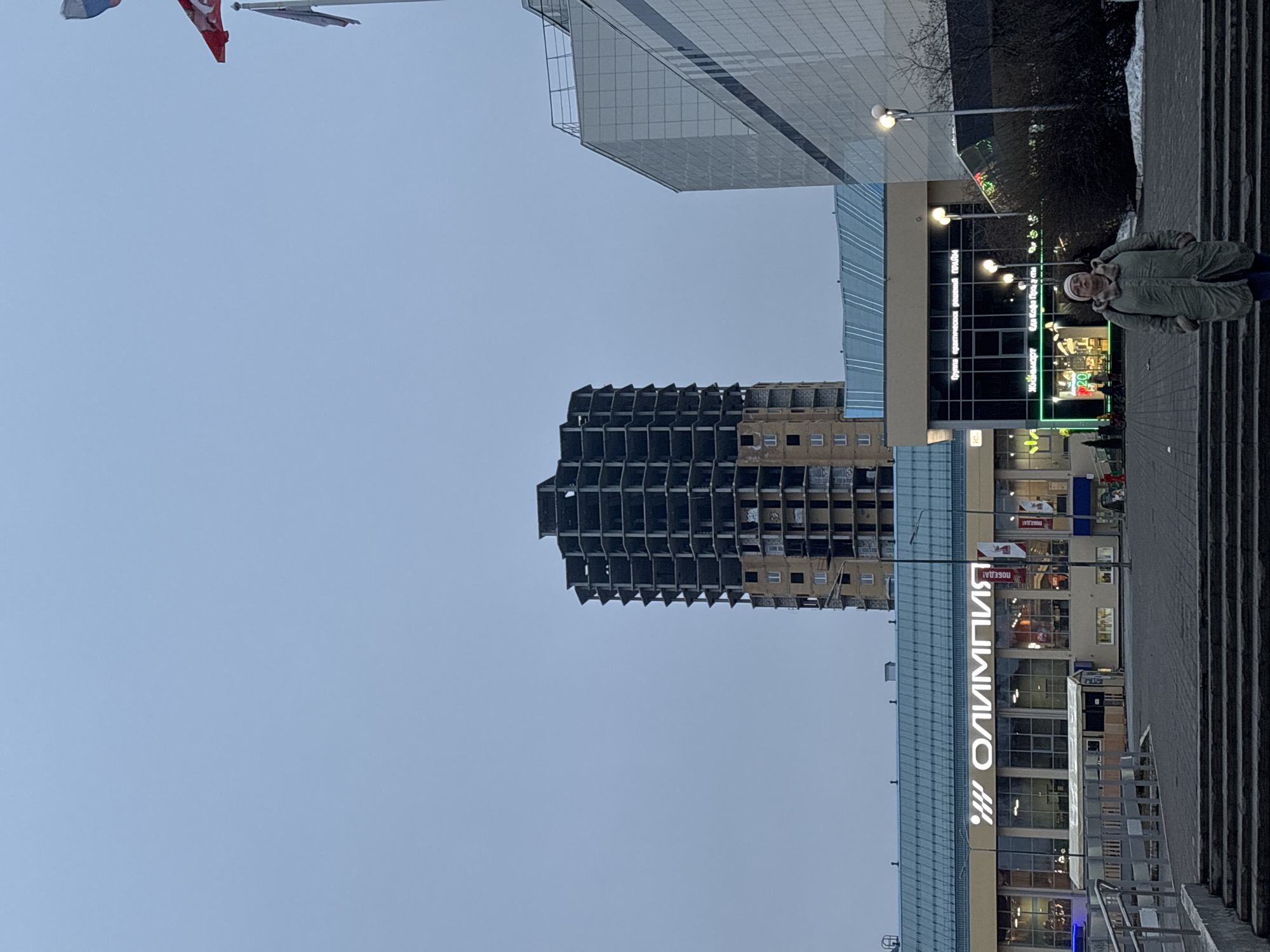 The image captures an urban scene in Perm, Russia. Prominently featured is the Olympia shopping center, easily identifiable by the large "ОЛИМПИЯ" sign. A partially constructed or derelict high-rise building stands nearby. A woman in a jacket is present in the scene. The environment appears to be a city street with steps leading up to the building. Flags are visible in the sky.