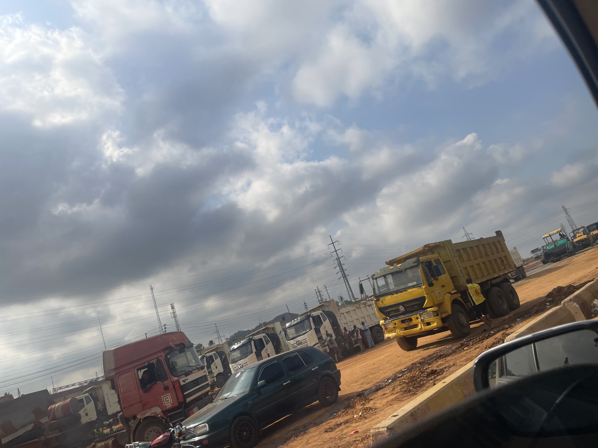 The scene appears to be a roadside area on the outskirts of Abuja, Nigeria. The location is a construction or industrial site, with an unpaved, reddish-brown dirt surface. Several large vehicles are present: A yellow dump truck is prominently featured, alongside multiple white dump trucks and a red truck. There is a dark green hatchback car parked near the red truck. Several people are visible standing near the white trucks, possibly engaging in conversation. Overhead, electrical transmission towers are visible against a cloudy sky. The image is likely taken from inside a car looking out onto the scene. The environment suggests ongoing road construction or heavy industrial activity.