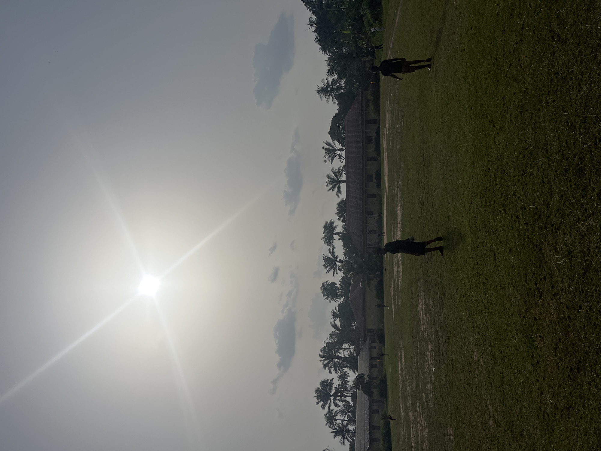 The image shows an outdoor scene, likely in Ikot Ekpene, Nigeria. Two individuals are present in the image, positioned in a field of grass. The background includes palm trees and a building with a visible roof. The sky is partially obscured by clouds, and the sun is bright.