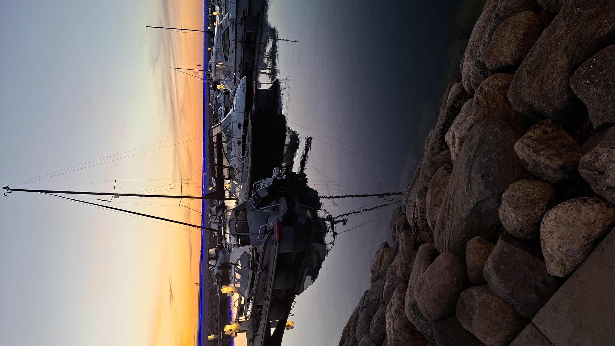 The image depicts a marina at sunset, with boats docked and their reflections visible in the calm water. The sky transitions from a vibrant orange and yellow near the horizon to a soft blue overhead. A sailboat with a tall mast stands prominently, with other smaller motorboats moored alongside. A rocky shoreline is visible in the foreground, with some of the rocks illuminated by the fading light. The scene is peaceful and serene, capturing the beauty of a coastal landscape at dusk. The specific location in Tallinn, Estonia, is not discernible from the image alone.