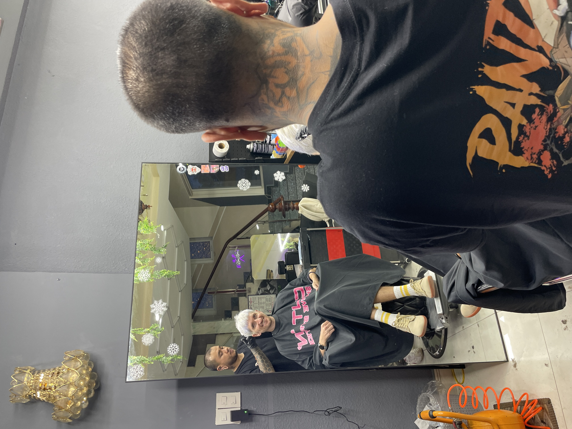 In a barbershop located in Xuân Hương - Đà Lạt, Vietnam, a person with short, spiky white hair is seated in a barber's chair, draped in a black cape with pink lettering. They are smiling and looking at themselves in a large, floor-to-ceiling mirror. Behind them, reflected in the mirror, a barber with short dark hair and tattoos on their arms and neck is in the process of cutting the customer's hair. The barbershop has grey walls, and a large ornate golden chandelier hangs from the ceiling. The mirror is decorated with hanging green vines and white snowflake cutouts. The floor is tiled and there are scattered hair clippings around the barber's chair. In the background, reflected in the mirror, a staircase with a wooden railing and neon lighting is visible. On a shelf to the right of the mirror, various hair products and a roll of paper towels are present. To the left of the mirror, a black electrical outlet is mounted on the wall.