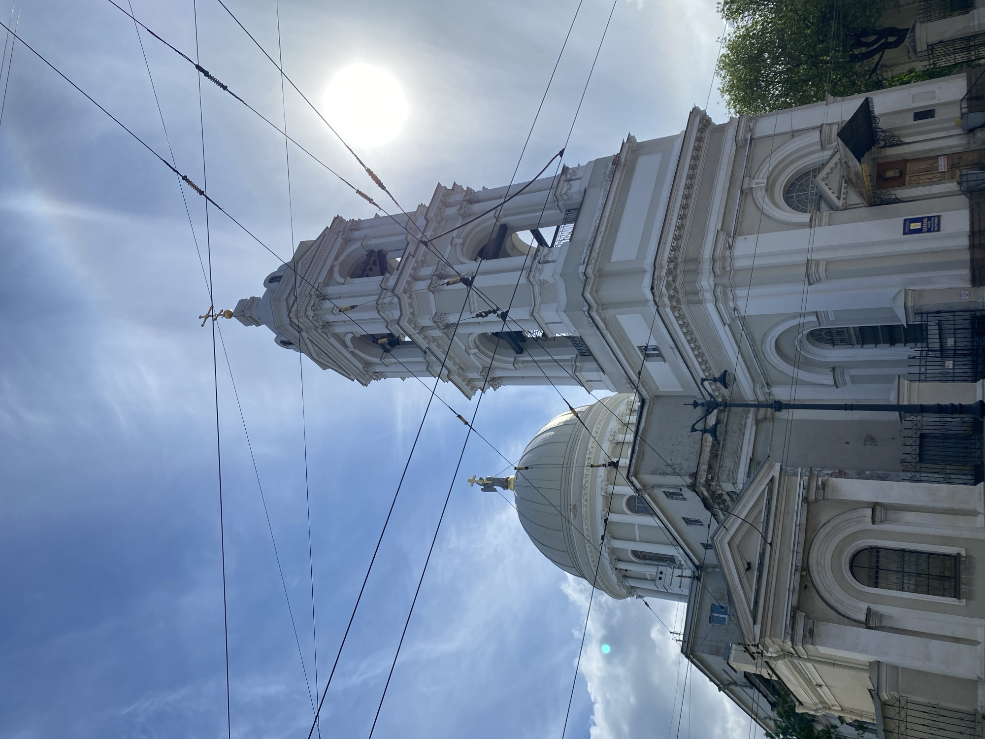 The image shows a view of St. Catherine Armenian Church in St. Petersburg, Russia. The building is white with a prominent bell tower and a dome topped with a golden statue. Power lines intersect the view of the sky, which is light blue with scattered clouds and a bright sun. The shot is taken from a low angle, emphasizing the height of the church structure. Part of the building's side facade is visible on the right.