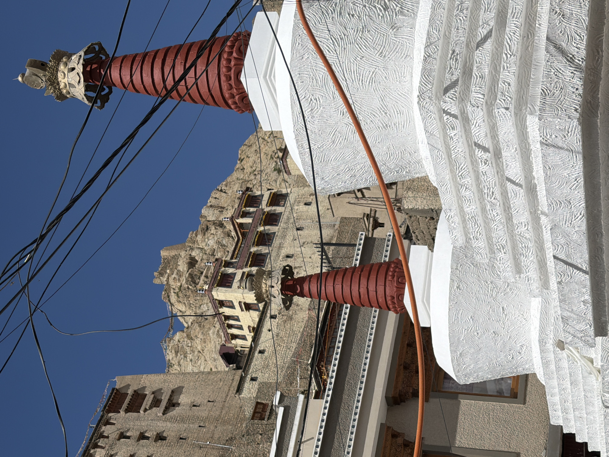 The image shows the stupas of Leh Palace in Leh, India. The stupas are white and adorned with red spires and golden finials. The palace itself is a multi-storied brick structure built into a mountainside. The sky is a clear blue, and power lines are visible crisscrossing the frame. The foreground features the stepped base of a large stupa, with its intricately textured white surface. In the background, another stupa, smaller and older, is partially visible. The overall scene conveys a sense of religious and architectural grandeur set against a dramatic natural landscape.