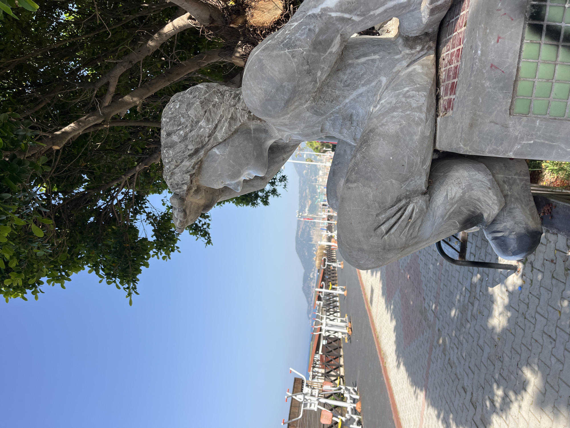 A slightly oblique full outdoor shot of a weathered grey stone statue of a woman looking pensive, with some modern-looking outdoor exercise equipment visible in the background.
The statue is the primary subject, displaying a simple style with a smooth face and wavy hair. She appears to be sitting or kneeling, with her arms crossed or clasped in her lap. A part of the statue's backside includes a pattern of red and green squares.
The background features the coastal landscape of Alanya, Turkey. The blue sky is partly obscured by tree branches. The foreground depicts a patterned brick walkway with exercise equipment placed along its side. A blurred mountain range can be seen in the distance, suggesting the statue is located near a beach or promenade. The lighting suggests a sunny day.