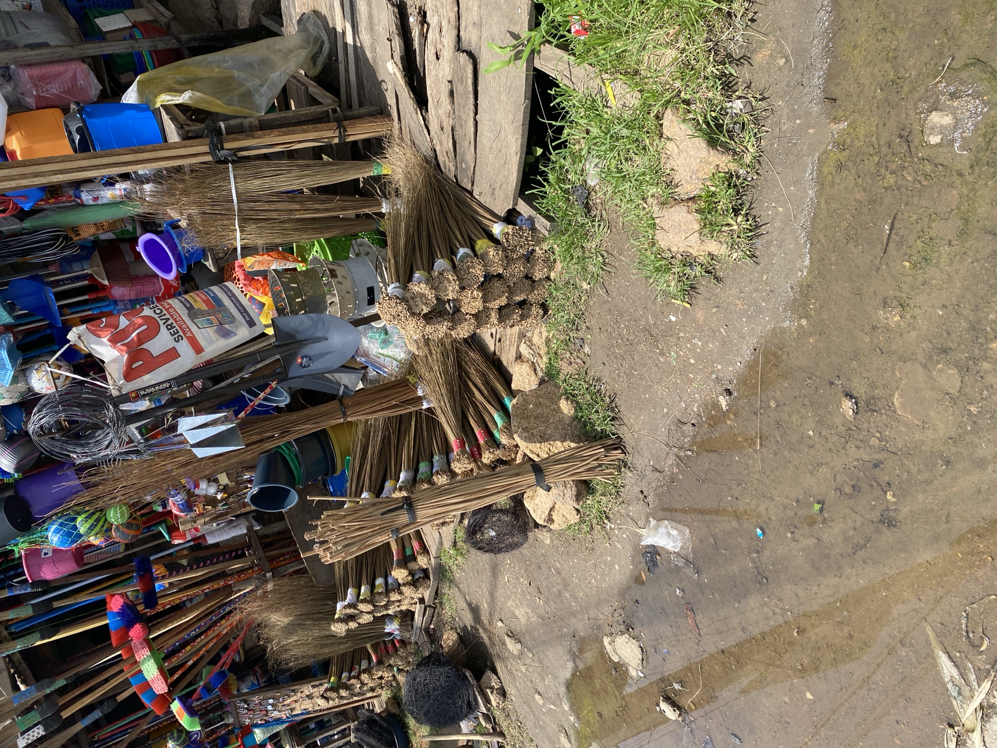 The image depicts an outdoor market stall in Jos, Nigeria, specializing in cleaning supplies, primarily brooms and shovels. The stall is situated on uneven, muddy ground with puddles of water, suggesting recent rain or poor drainage. The merchandise is densely packed, with numerous brooms of various sizes and designs leaning against each other and stacked in bundles. Some brooms have colorful bristles, while others are made of natural fibers. Several shovels with metal heads and wooden handles are also visible.
In the background, a variety of household items, including buckets, containers, and possibly other cleaning tools, are displayed. The overall impression is one of a busy, somewhat disorganized retail environment, typical of informal markets. The lighting suggests it is daytime, with bright sunlight casting shadows. The presence of natural vegetation, such as grass growing between rocks and dirt, indicates an area that is not fully paved. There are no people visible in the image, only the goods for sale and the surrounding environment.
