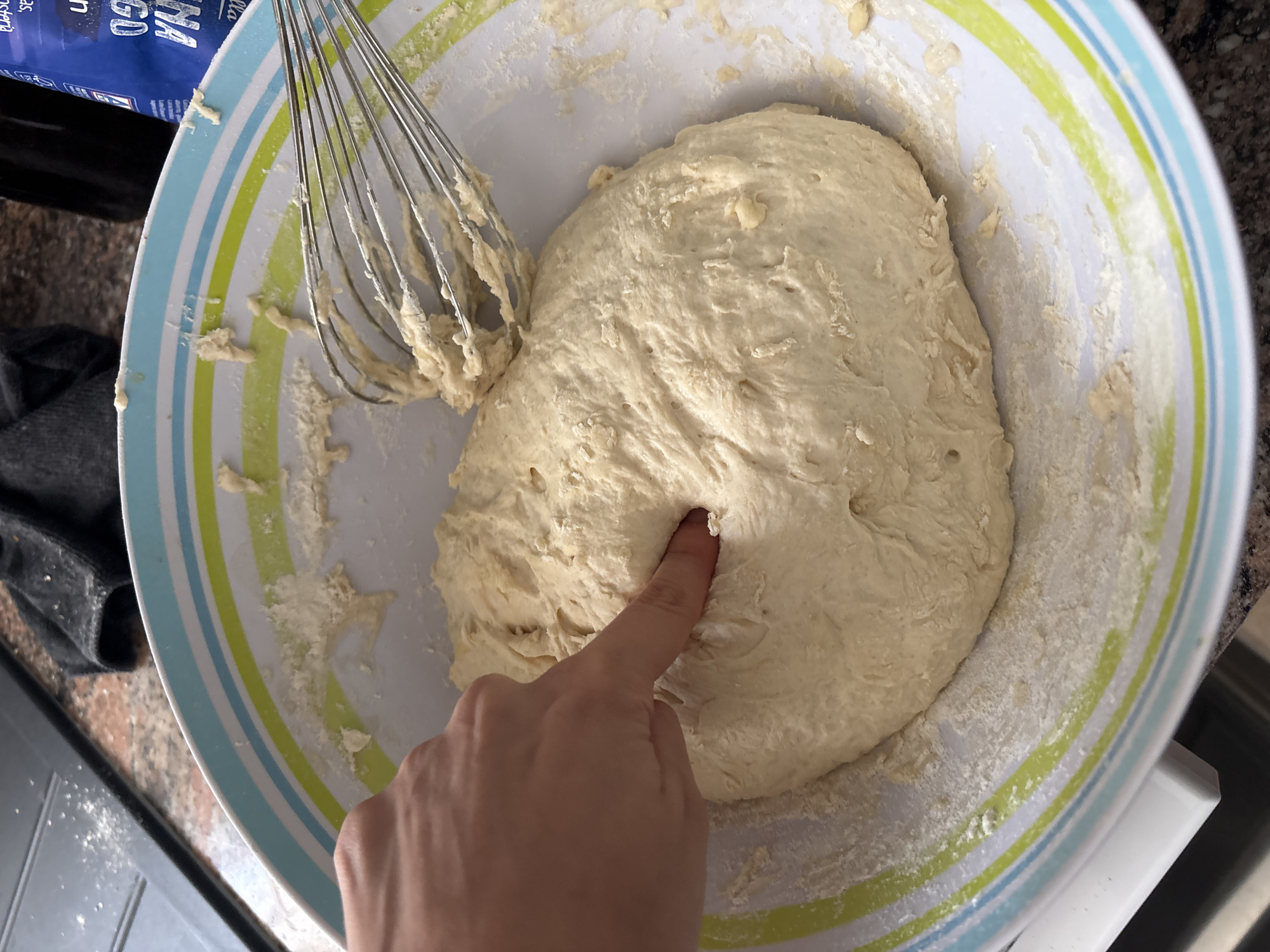 The image shows a close-up view of a person's hand pressing into a large ball of raw dough in a white bowl with blue and green stripes. A metal whisk is also visible in the bowl, with some dough clinging to its wires. The dough appears light in color and slightly sticky, with a few small air bubbles visible on its surface. Flour is dusted around the edges of the bowl and on the counter surface. In the upper left corner of the image, a portion of a blue container with white text is visible. The text on the container appears to be in a foreign language, with some discernible letters including "NA GO." The setting appears to be a kitchen, with a granite countertop visible in the background. The lighting suggests it is daytime. The activity taking place is likely the preparation of dough for baking, possibly bread or pizza, as indicated by the finger pressing into the dough, a common test for readiness or simply a way to interact with the dough during preparation. The overall impression is one of domestic activity, possibly a home baker at work. The location is specified as Orihuela, Spain, which does not provide any further visual cues within the image itself.